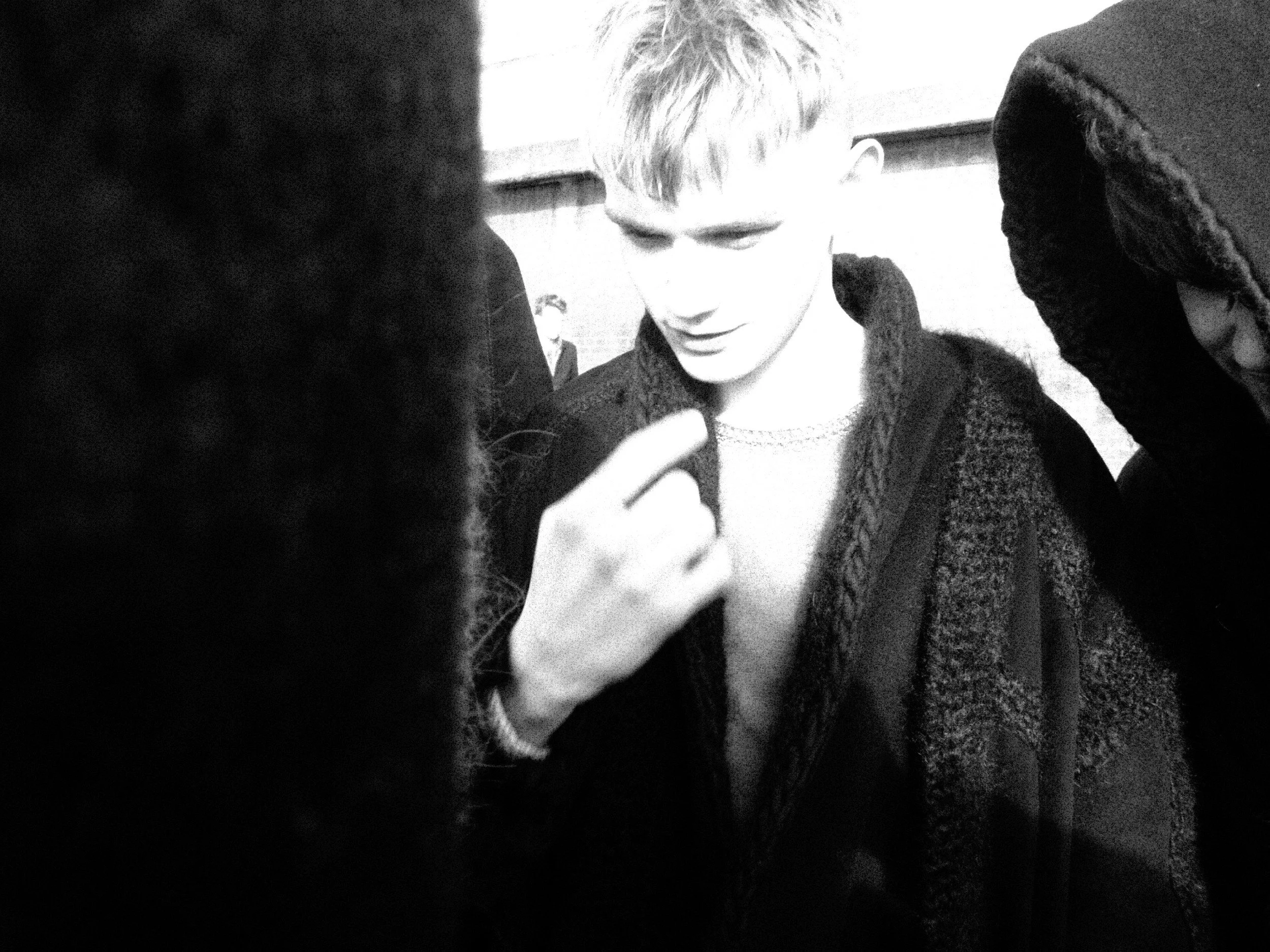 A black and white photo showing a young man with short, light hair looking downwards, surrounded by people. There are blurred figures in the foreground, and a person with dark, curly hair near the right edge of the image.