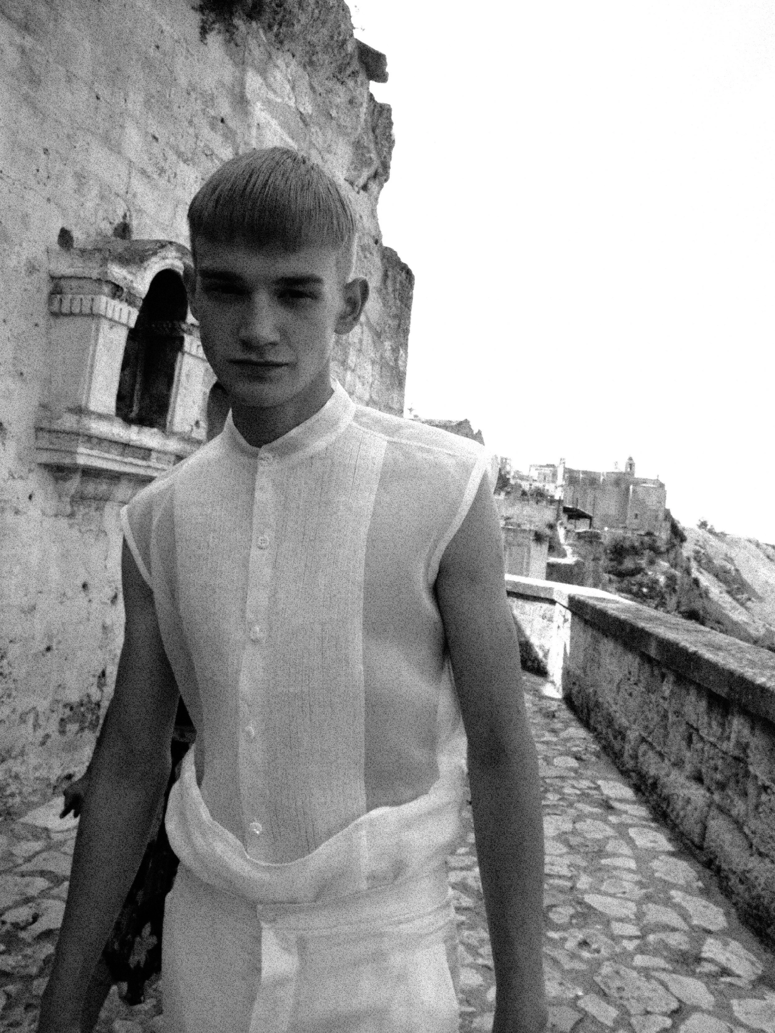 A young man with short hair and wearing a traditional style shirt standing outdoors on a cobblestone street with stone buildings in the background.