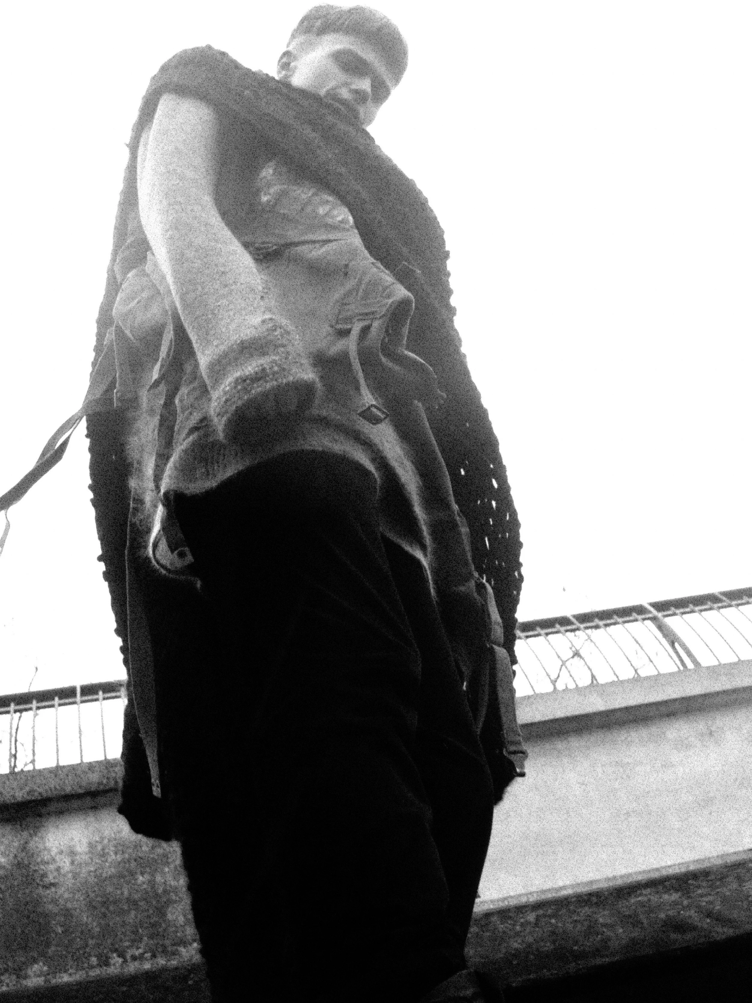 Black and white photo of a person standing outdoors near a railing, wearing a hoodie and jeans, looking down.