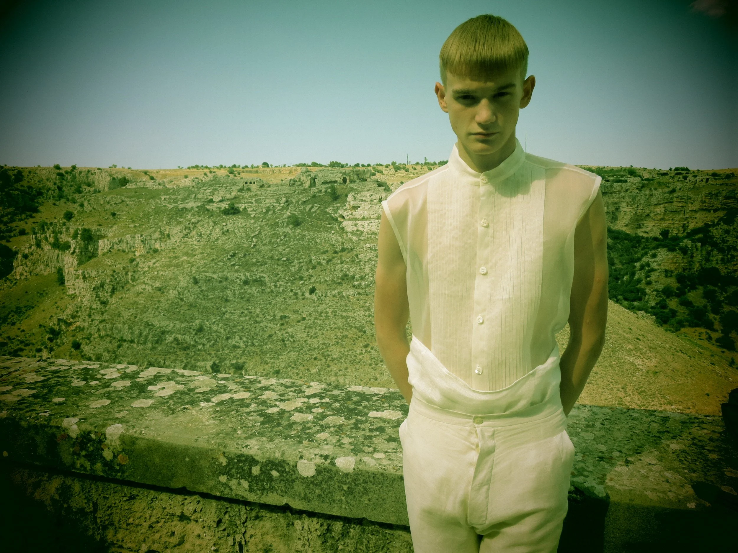 A young man with light skin and short blond hair stands outdoors in front of a rocky, grassy landscape with sparse trees under a clear blue sky. He is wearing a sleeveless white shirt and white pants, with his hands in his pockets, looking at the cam