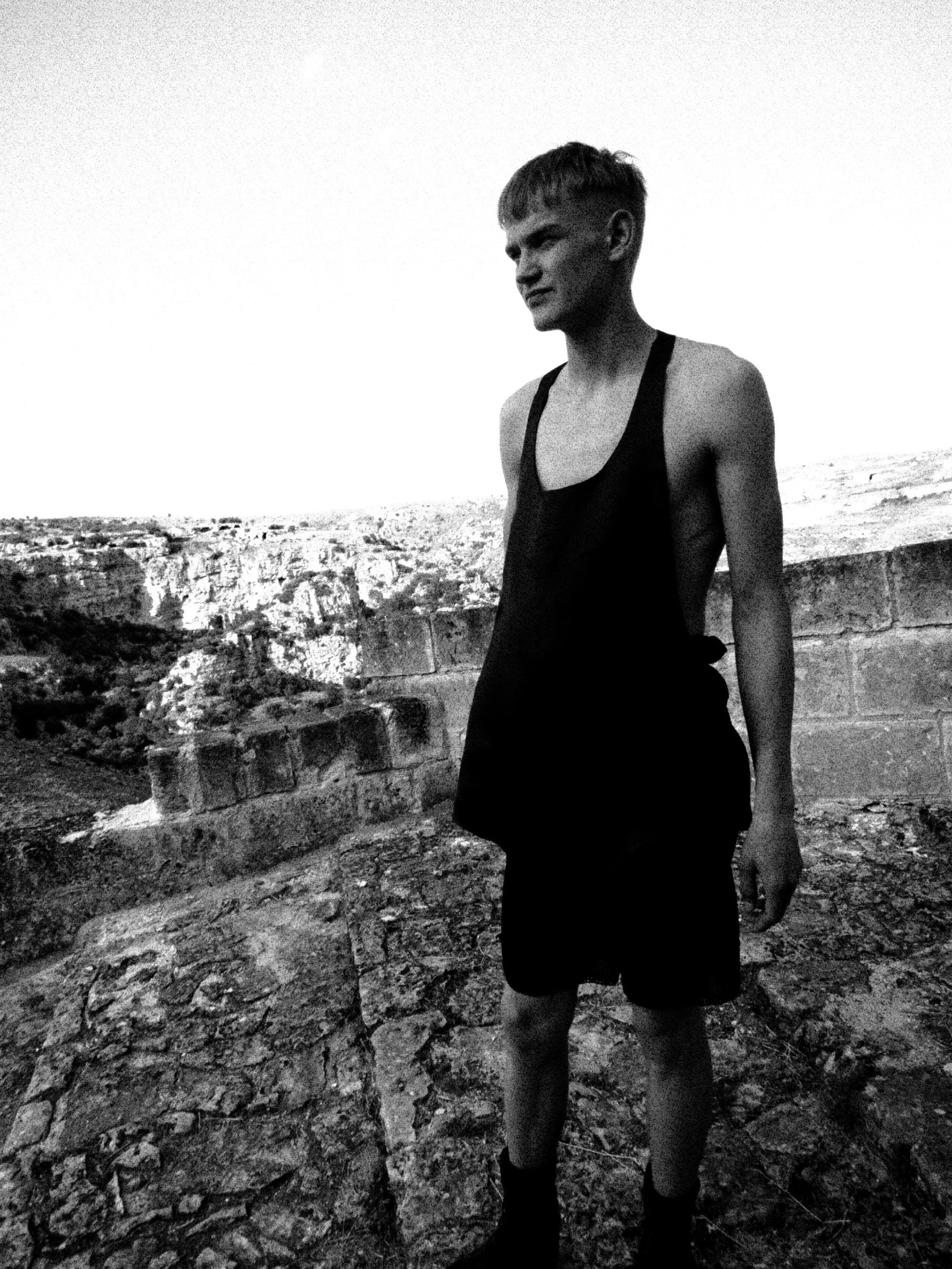 A young woman with short hair wearing a sleeveless top, shorts, and knee-high boots, standing near a stone wall outdoors with a landscape of cliffs and rocky terrain in the background, in black and white.