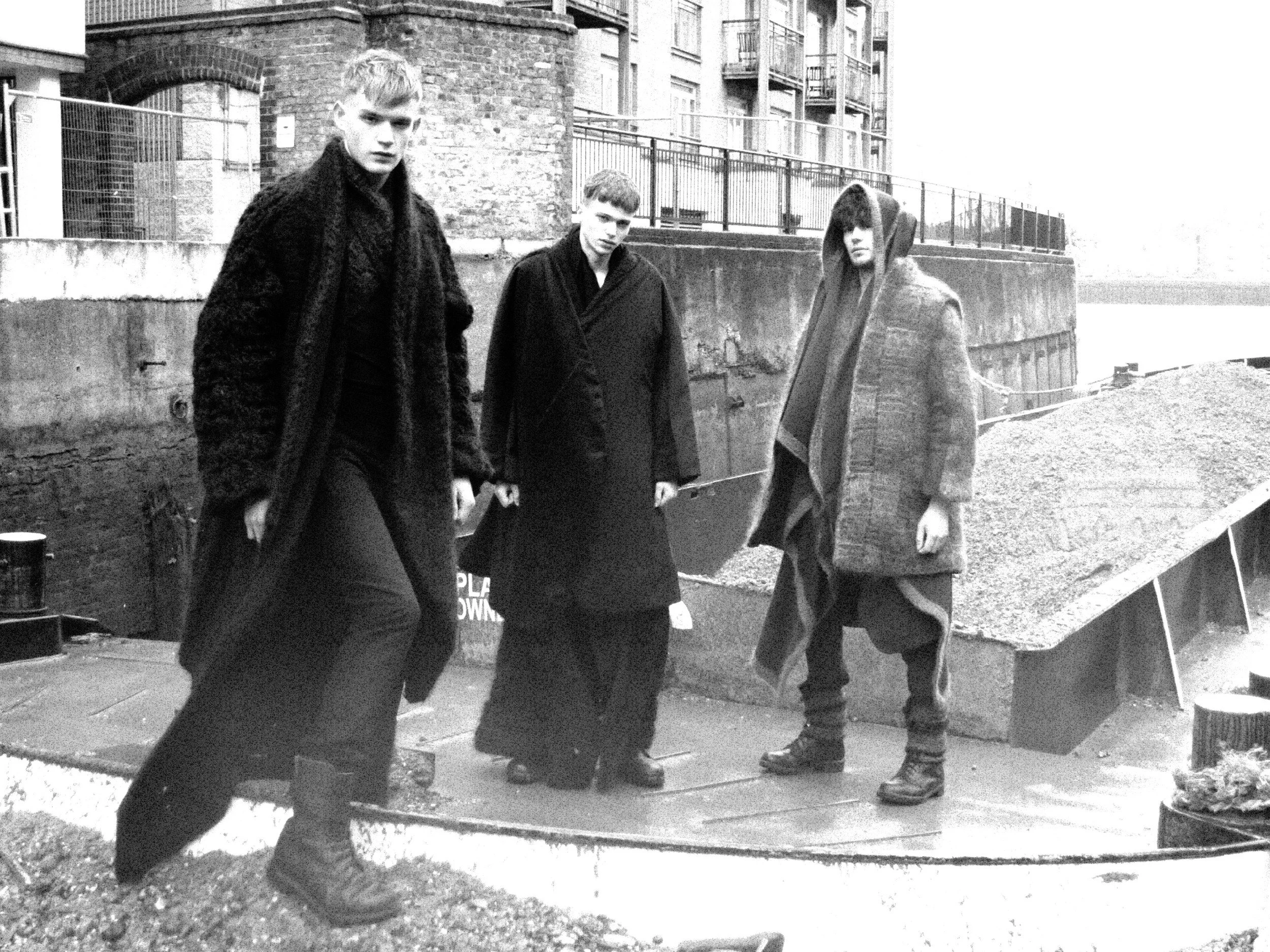Three young men in dark clothing walking along a sidewalk in an urban area with brick buildings and balconies behind them.