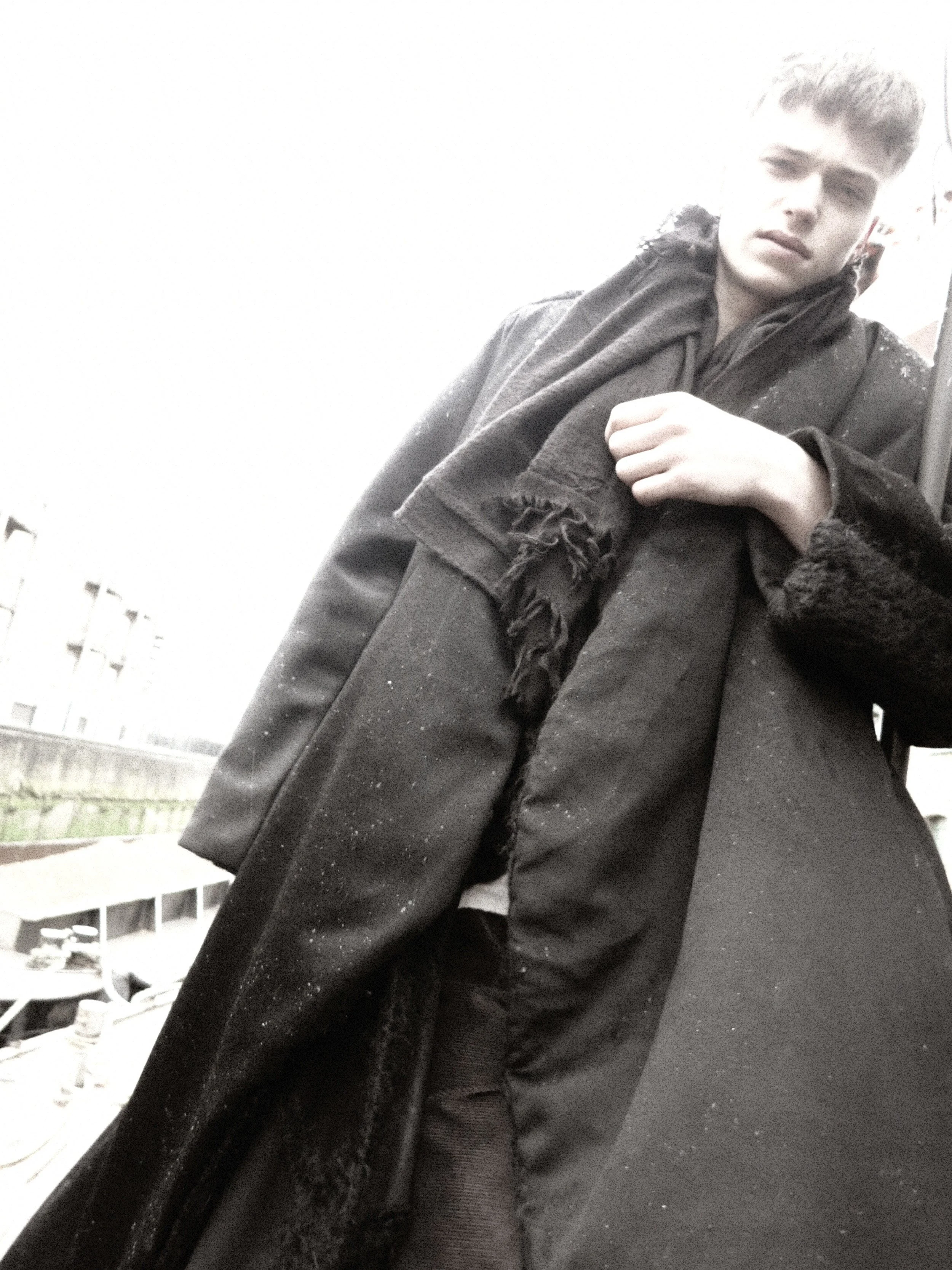 A young person with short hair, wearing a dark coat and scarf, standing outdoors with their hand on their chest, near some buildings and a fence.