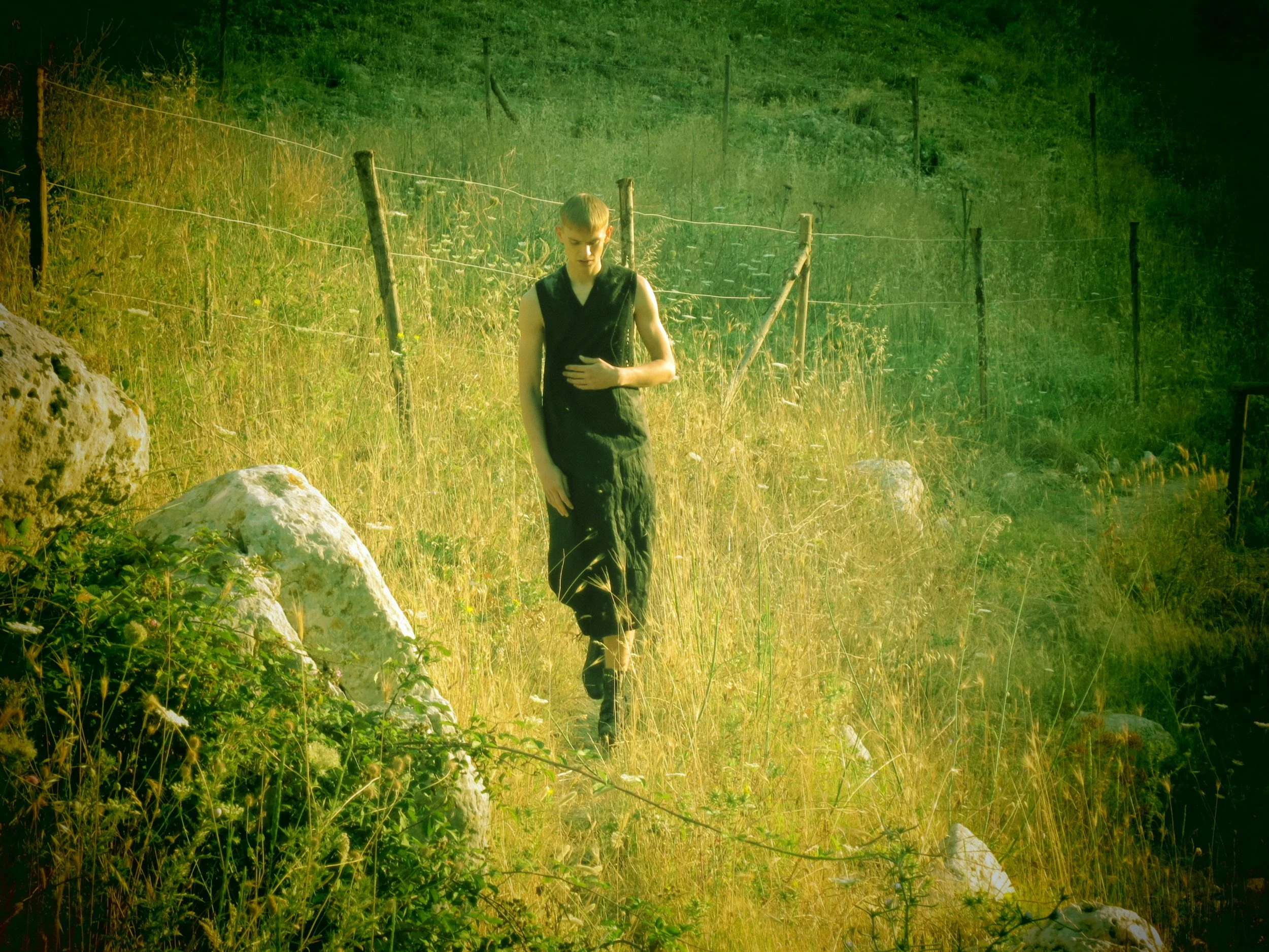 A young woman in a black sleeveless dress walking along a dirt path in a grassy, hilly outdoor area with a fence line on her right.