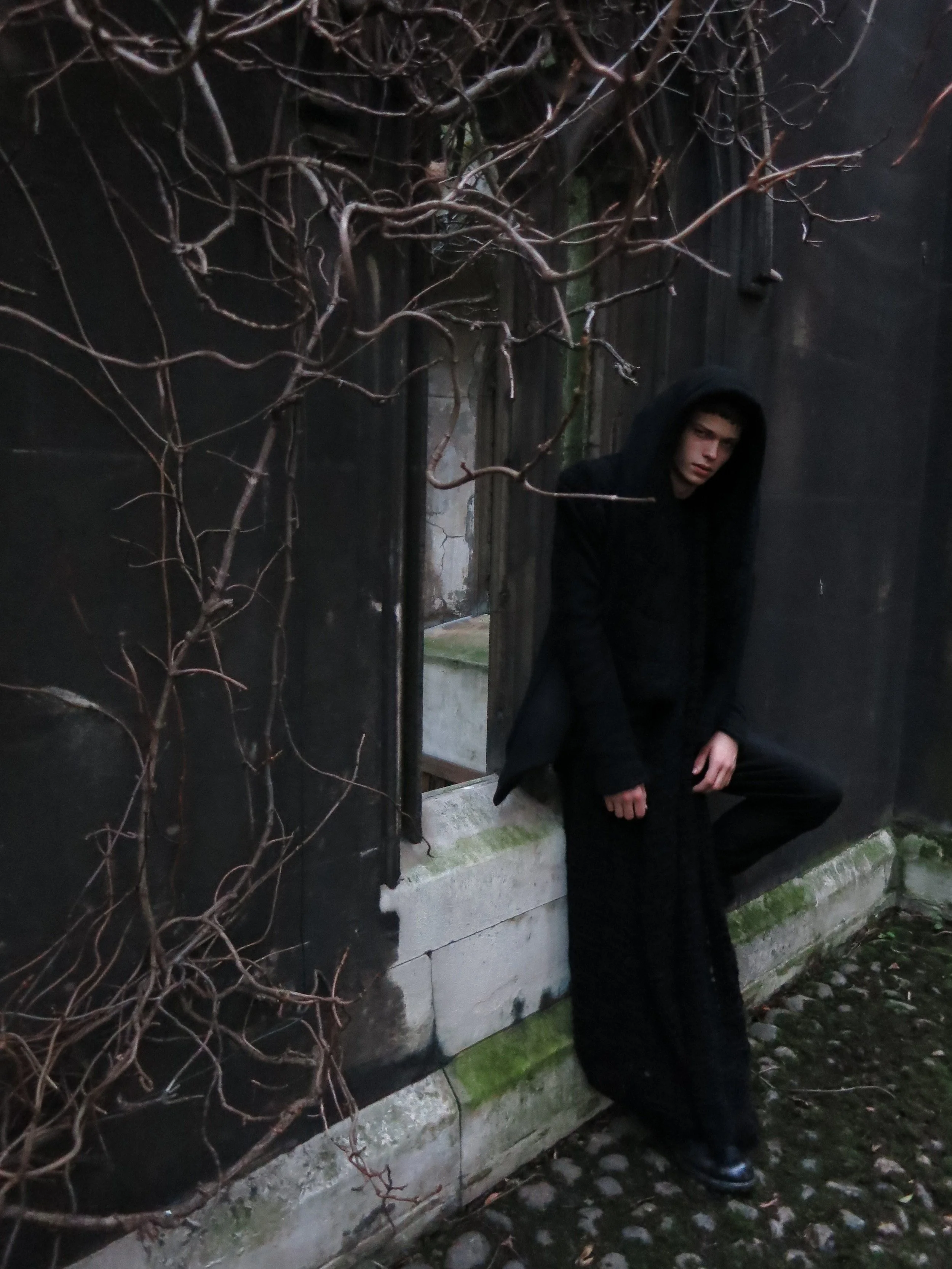 A young man wearing a black hoodie and black pants leaning against a black fence with leafless vines, in an outdoor setting with cobblestone ground.