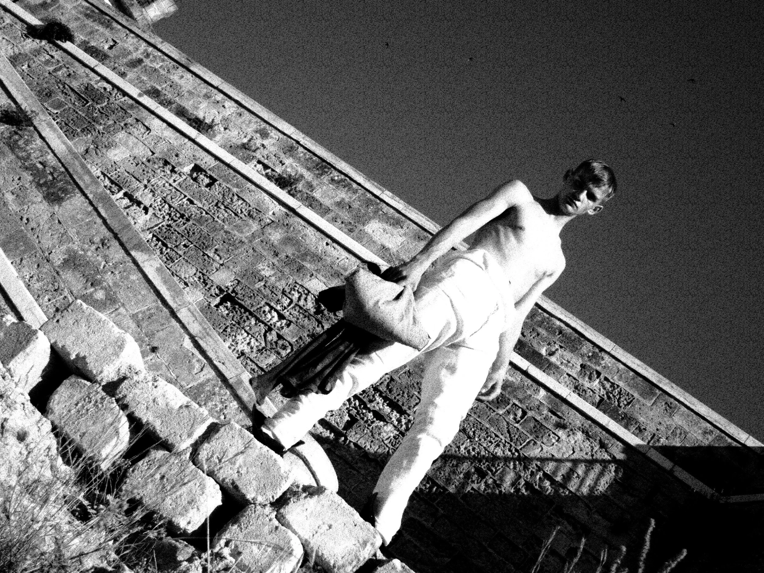 Black and white photo of a boy sitting on a stone ledge outdoors next to a brick wall at night.