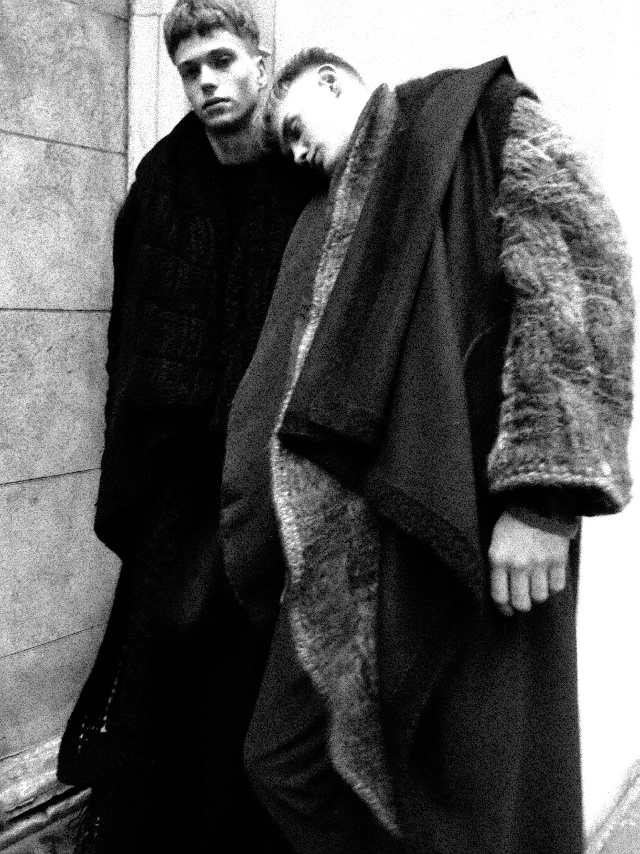 Two young men with short hair, wearing dark and textured coats, leaning against a wall in black and white.  Marc Lafia fashion photography