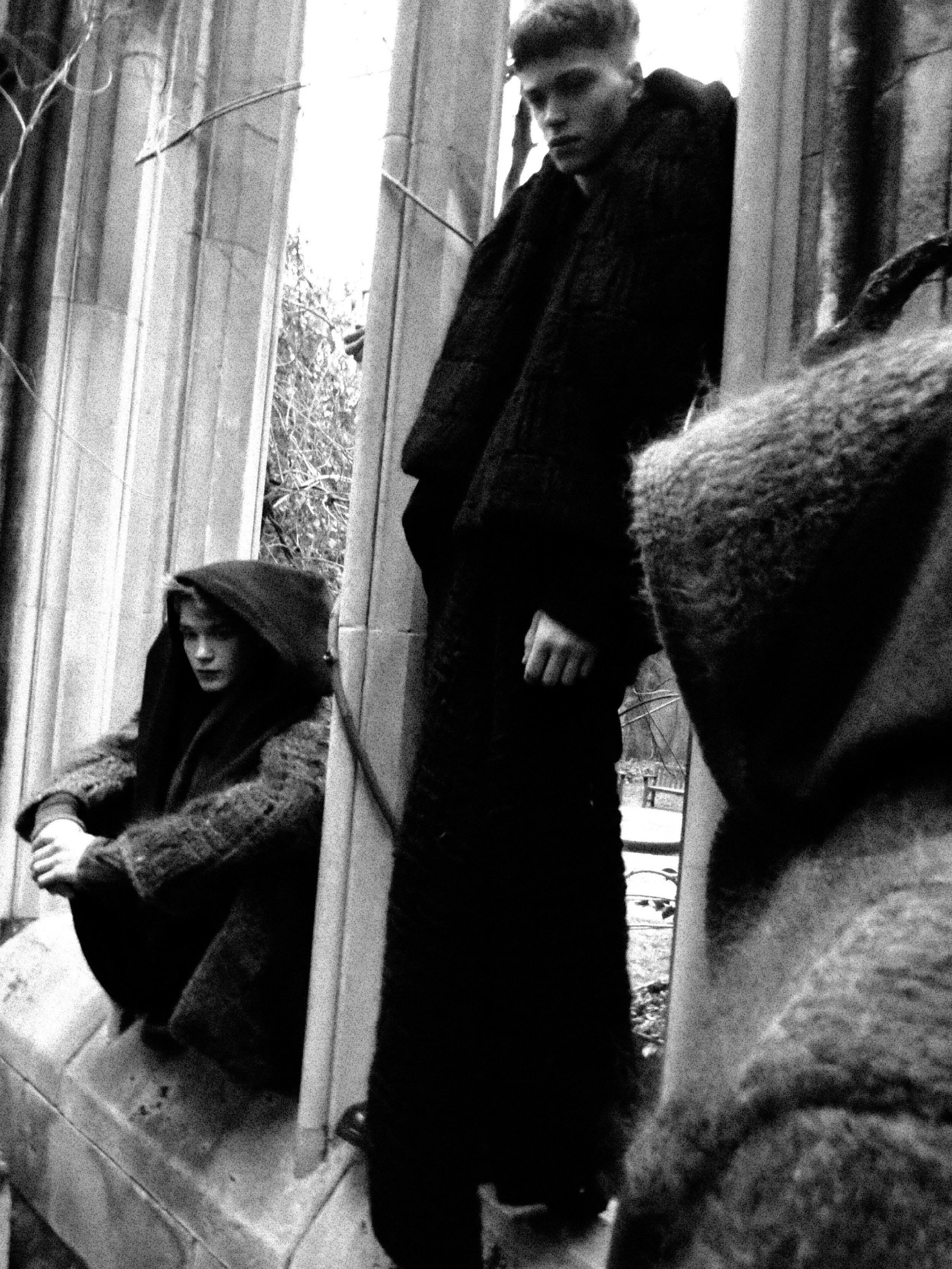 A black and white photo of three young men standing and sitting outside, near a building with large windows. One young man is sitting with a hood over his head, looking slightly down, while the other two are standing, with one looking at the camera a