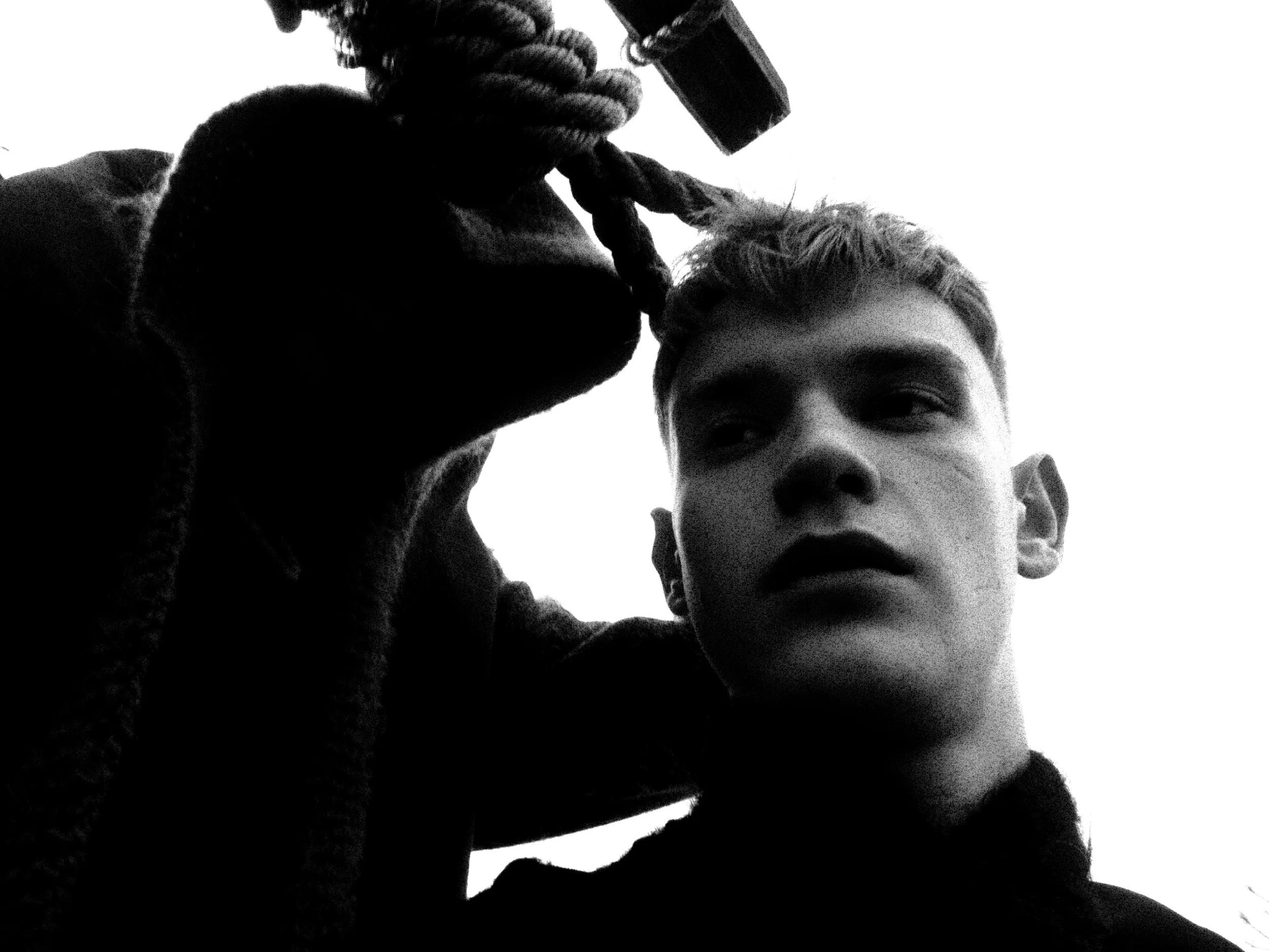A person with short hair wearing a hoodie, holding a rope or cable with one hand, and looking into the camera with a serious expression, in black and white.