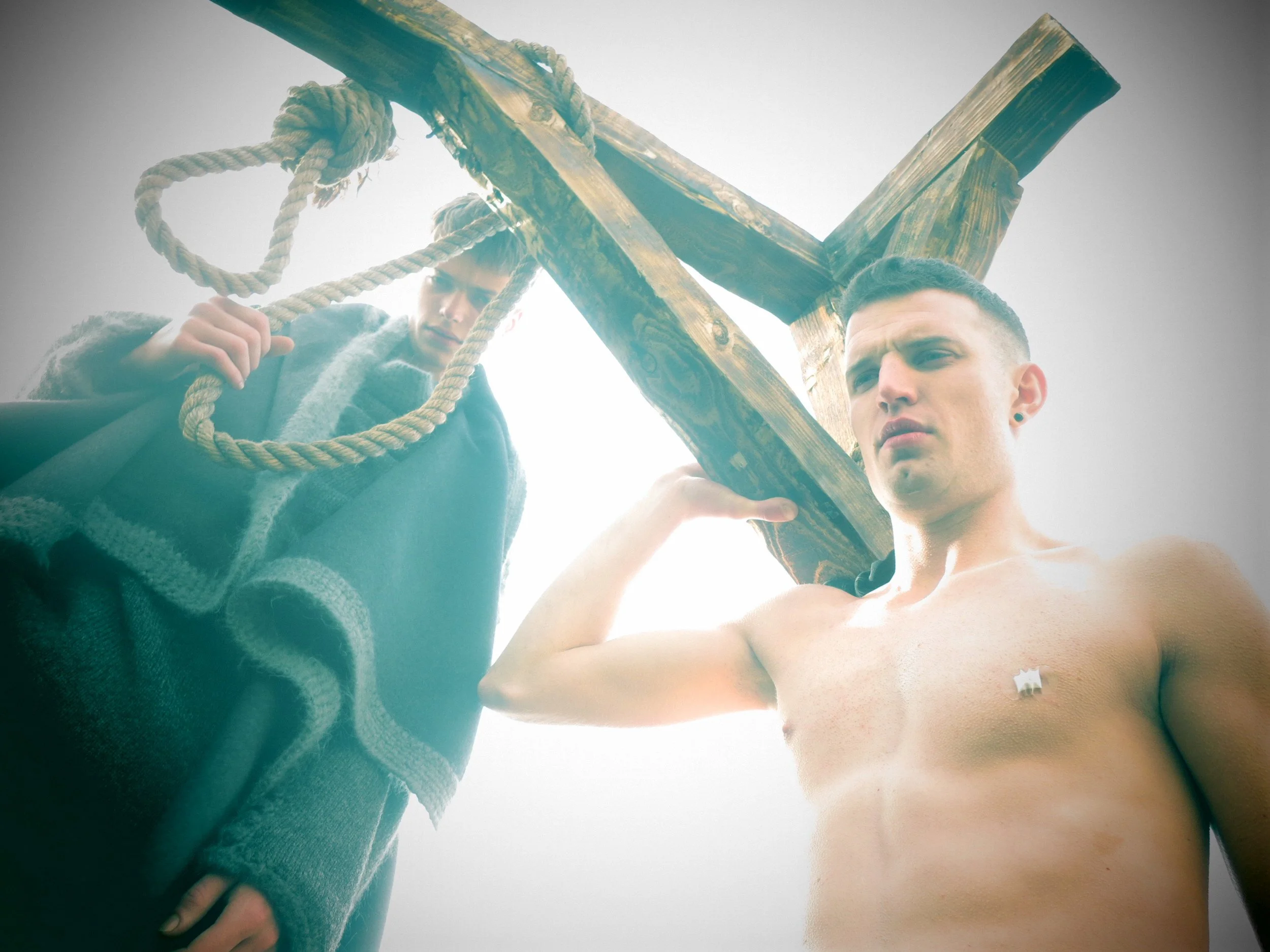 Two young men outdoors, one holding a wooden structure with ropes, both looking down at the camera, with a bright sky background.