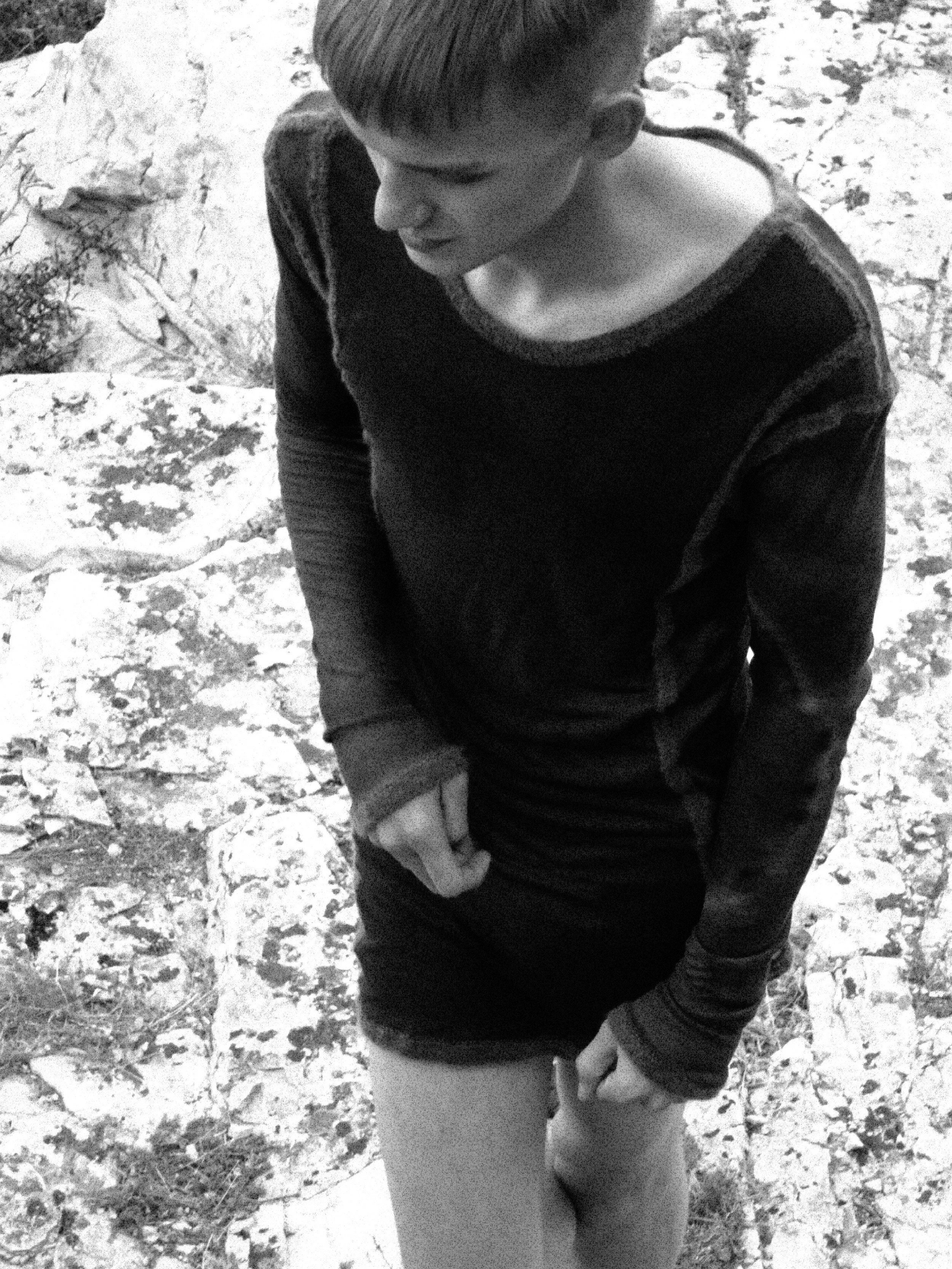 A young boy kneeling on a rocky surface, looking down with his hands resting on his knee, wearing a dark long-sleeve shirt, with short hair and a side-swept hairstyle.