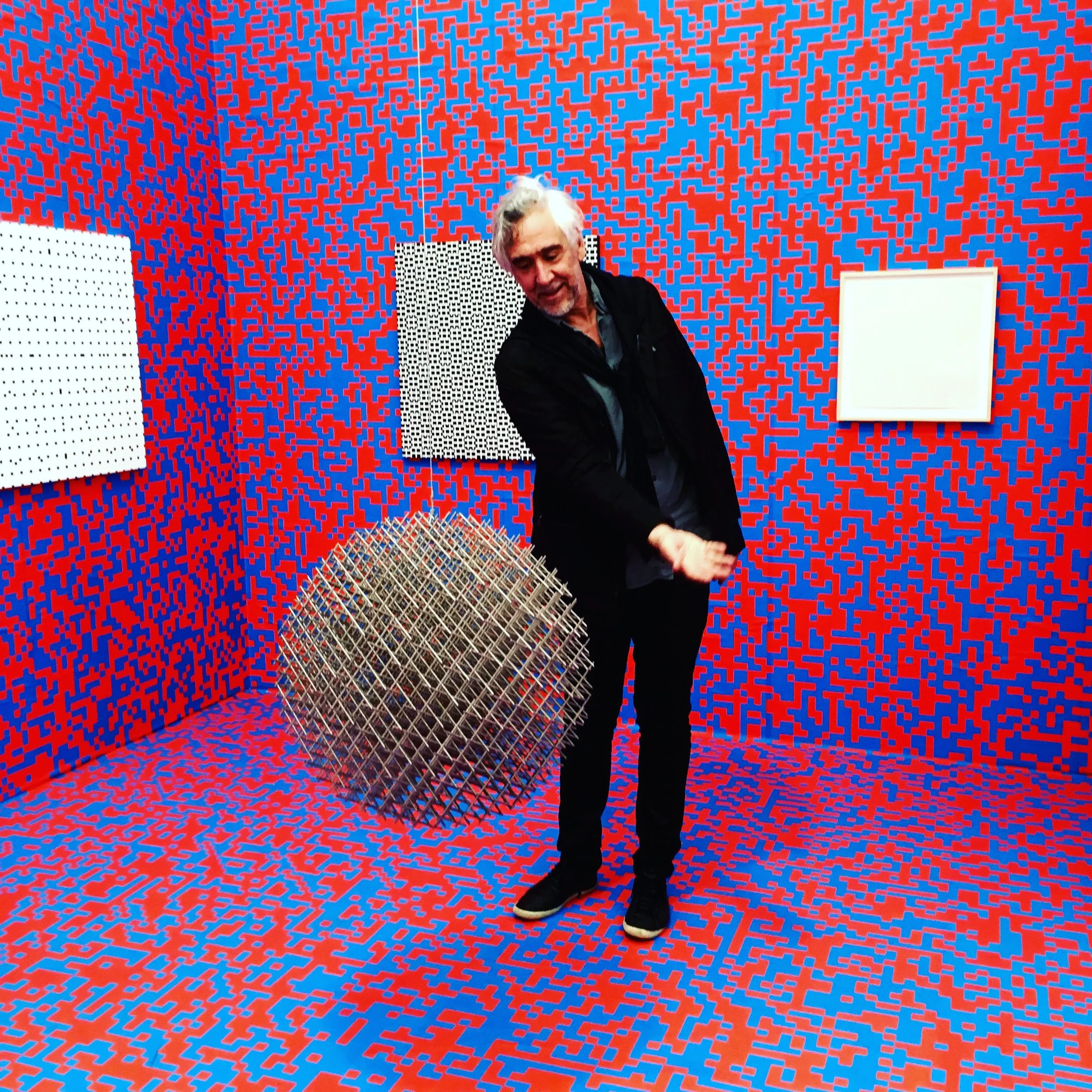 Man in black jacket with gray hair standing in an art gallery with vibrant red and blue pixelated patterned walls and floor, posing near a large metallic spherical sculpture and three pixelated art pieces on the wall.