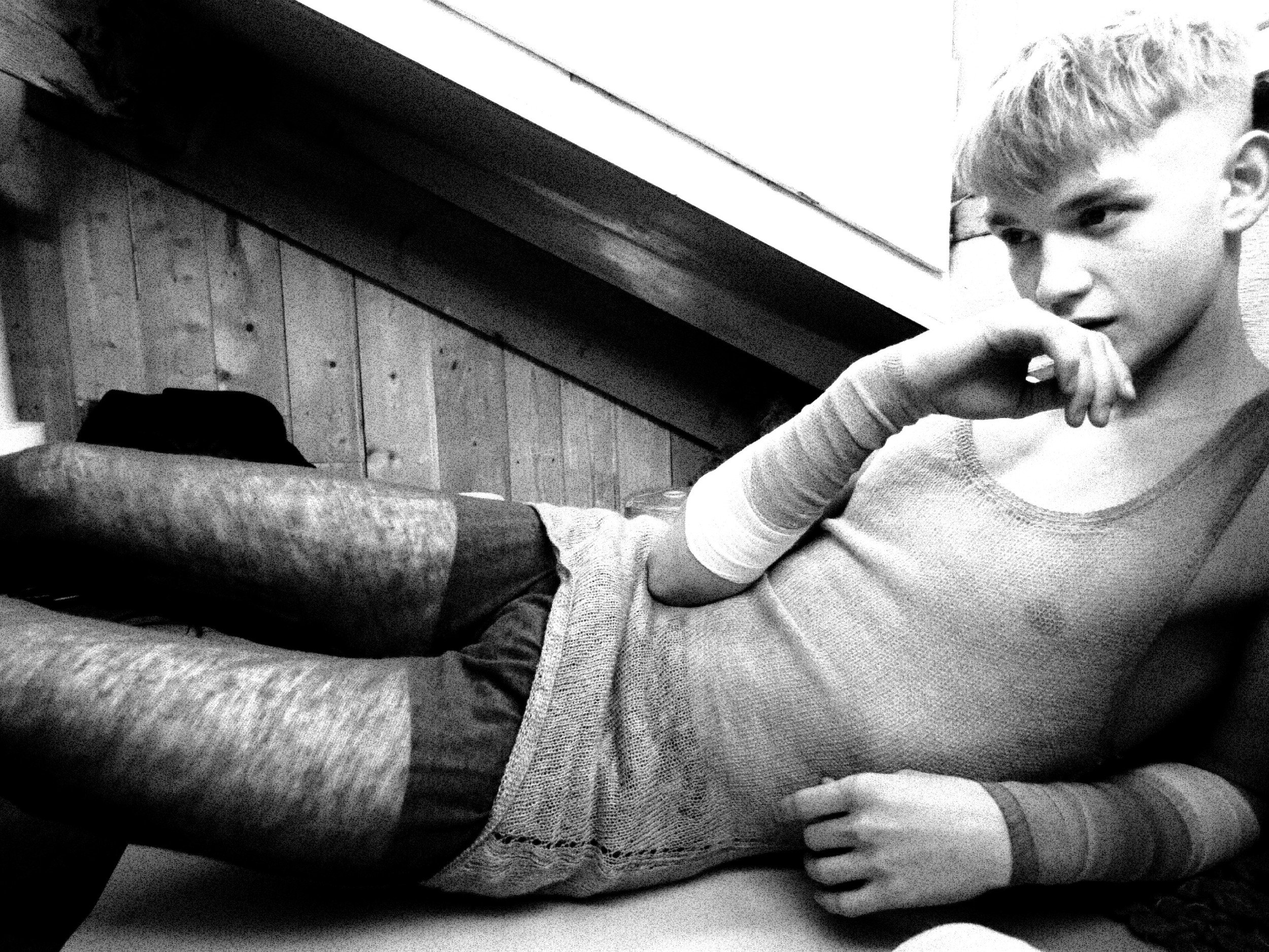 A black-and-white photo of a young man with light-colored hair, sitting on a cushioned surface, with his hand near his face, looking intently to the side.