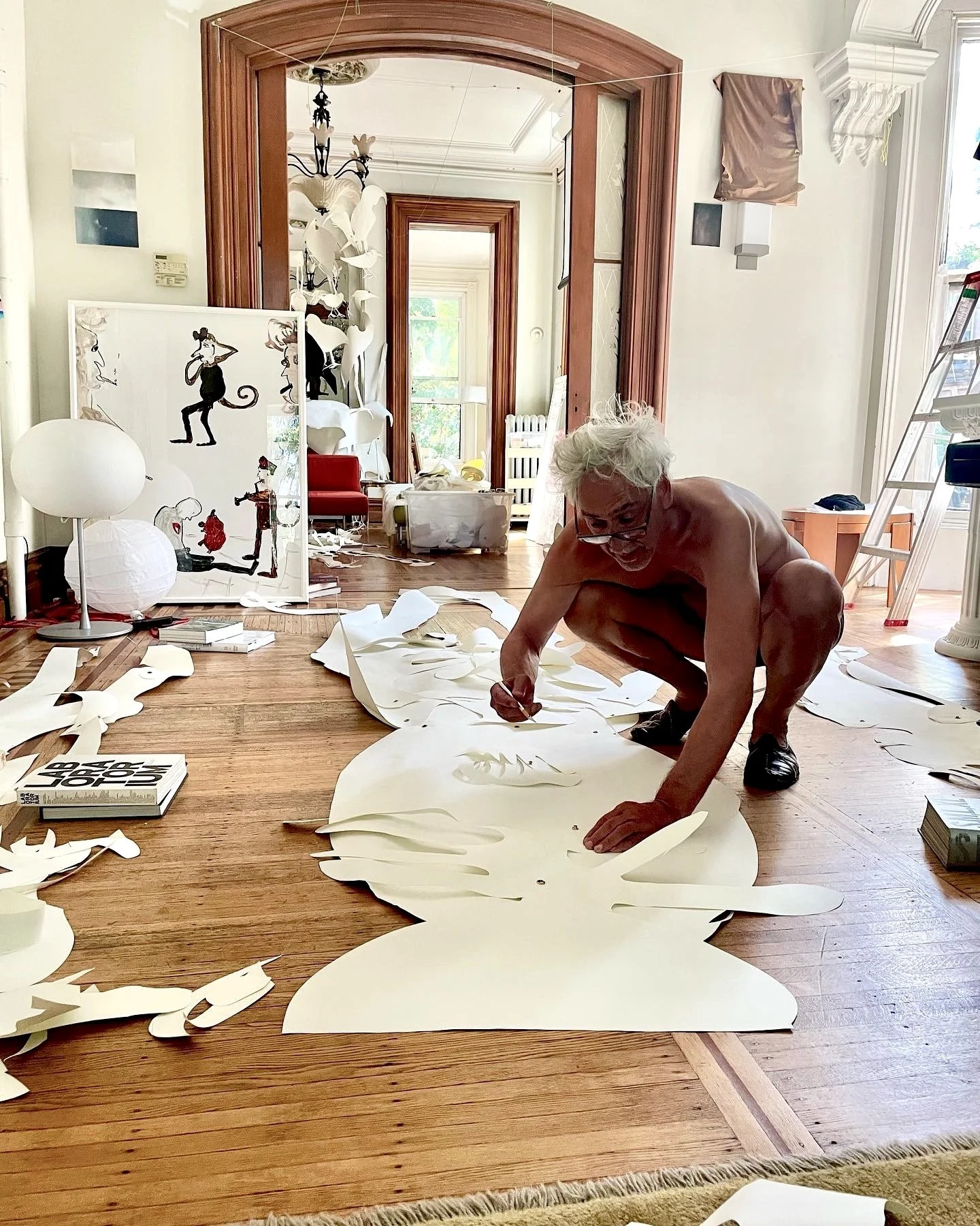 An elderly man with white hair and glasses squats on a wooden floor, working on large white paper cutouts. The room is filled with art supplies, papers, and books, with a background of a bright, spacious interior and artwork leaning against the wall.