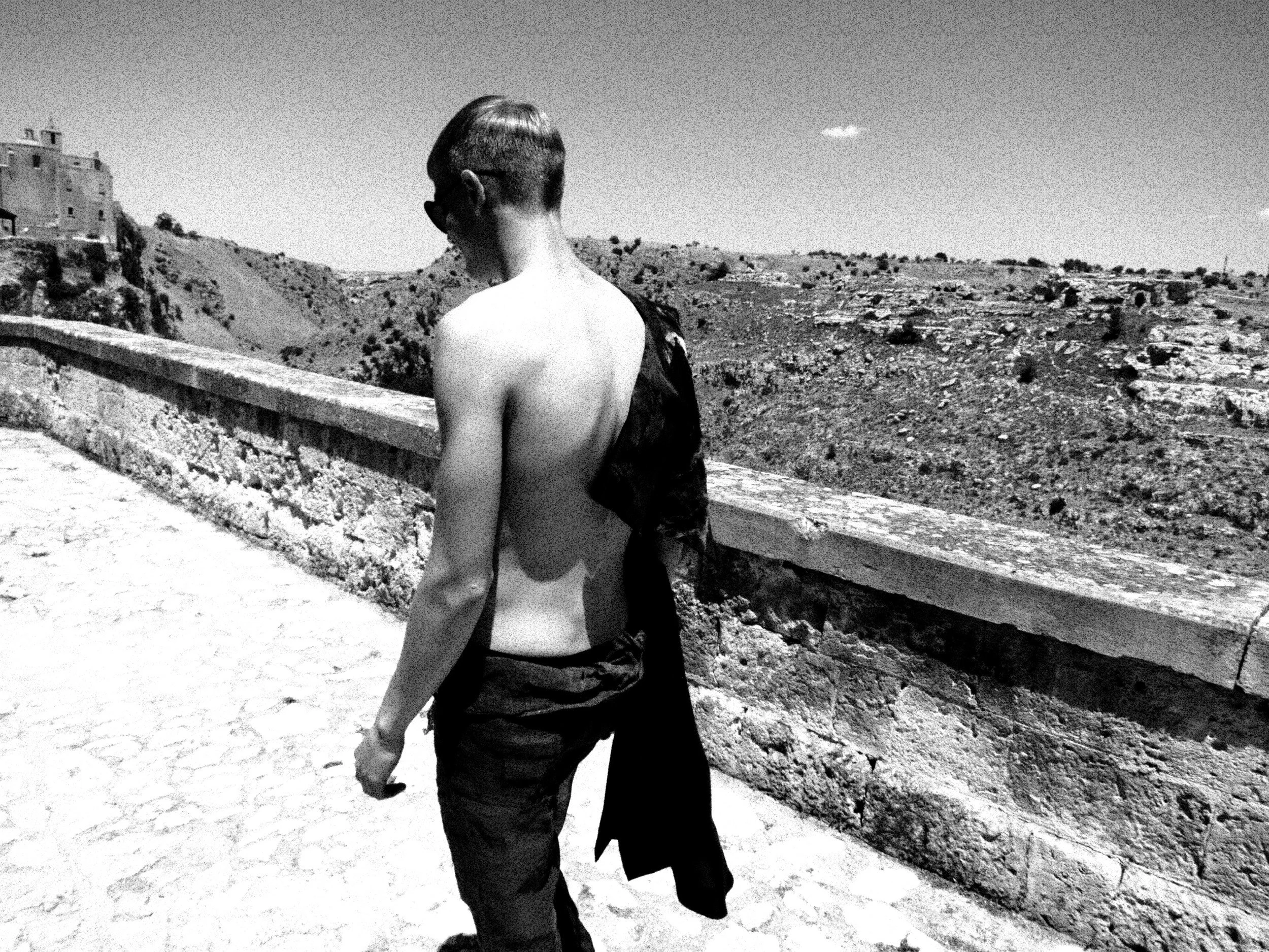 Black and white photo of a shirtless man with glasses walking on a stone path next to a stone wall, overlooking a hillside with sparse vegetation and a few buildings in the distance under a clear sky.
