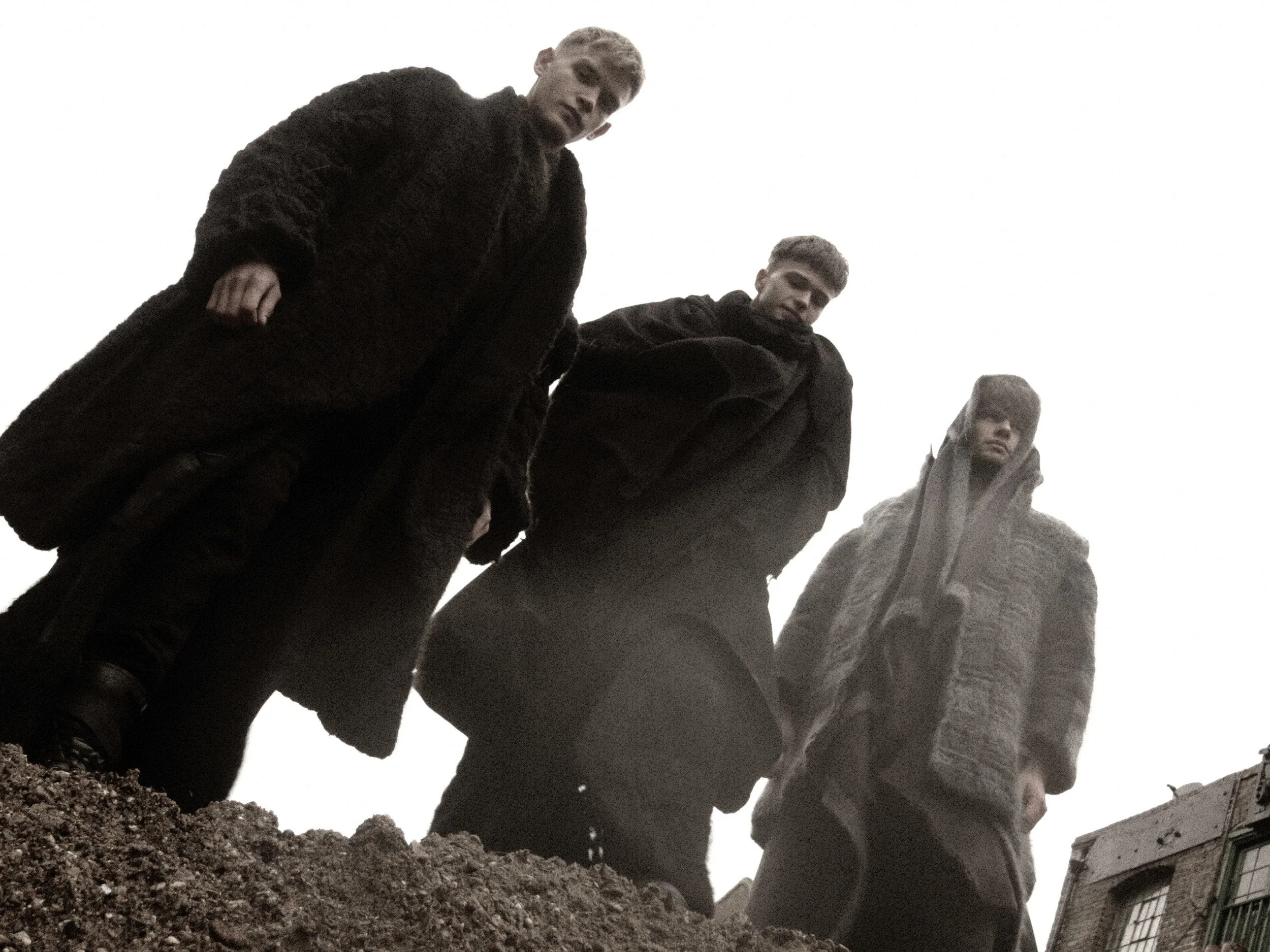 Three men standing outdoors, looking down at the camera, with a building partially visible in the background, and a cloudy sky overhead.