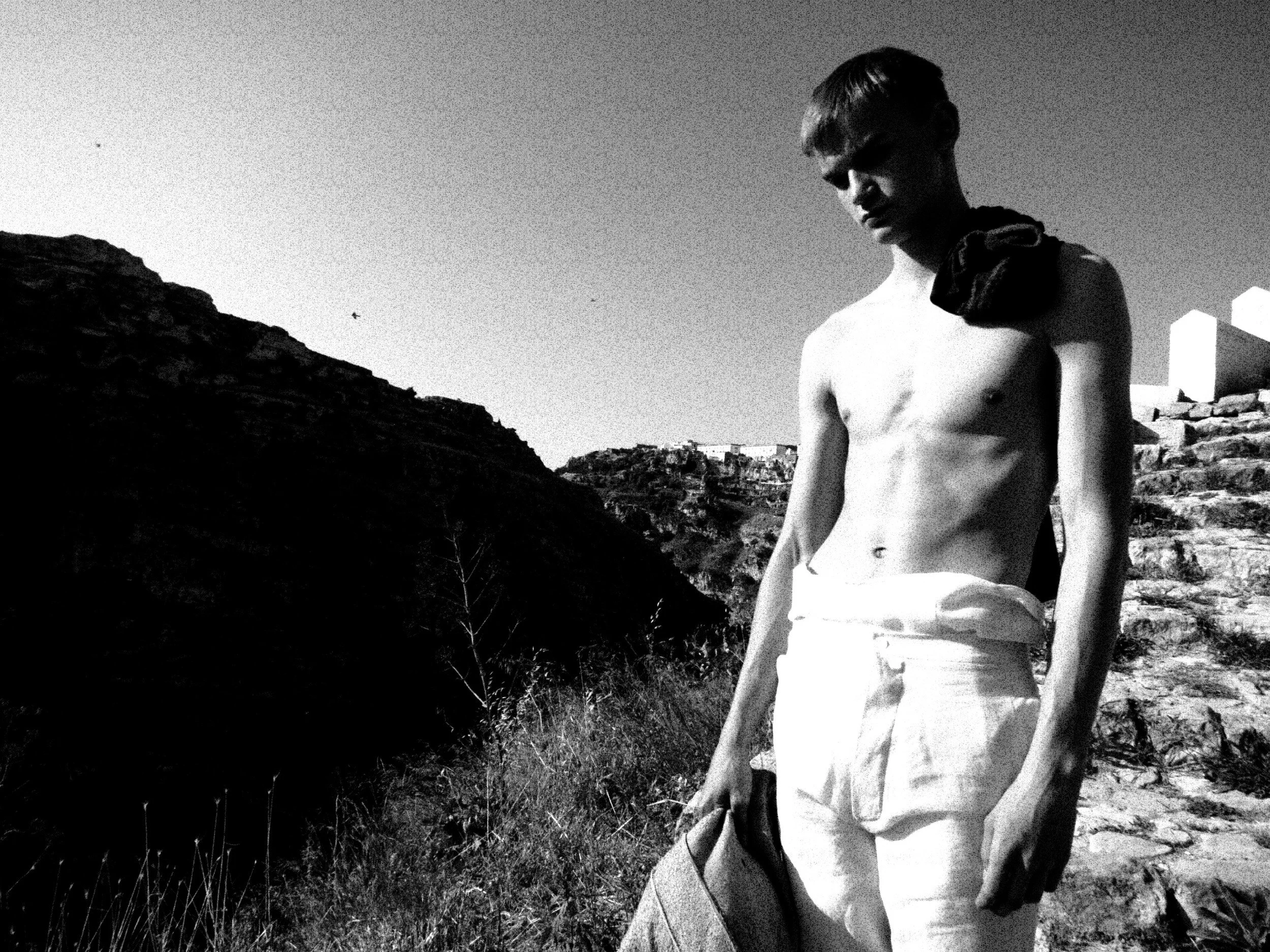 A black and white photo of a shirtless young man with short hair, wearing sunglasses and a scarf around his neck, standing outdoors on a rocky terrain with cliffs and a building in the background.