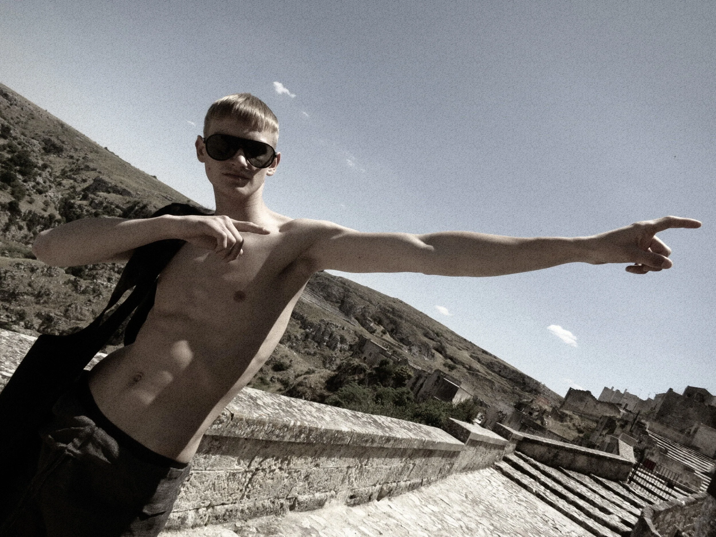 A shirtless young man wearing large black sunglasses extending his arm with fingers pointed outward, standing outdoors in front of hills and a cityscape under a partly cloudy sky.