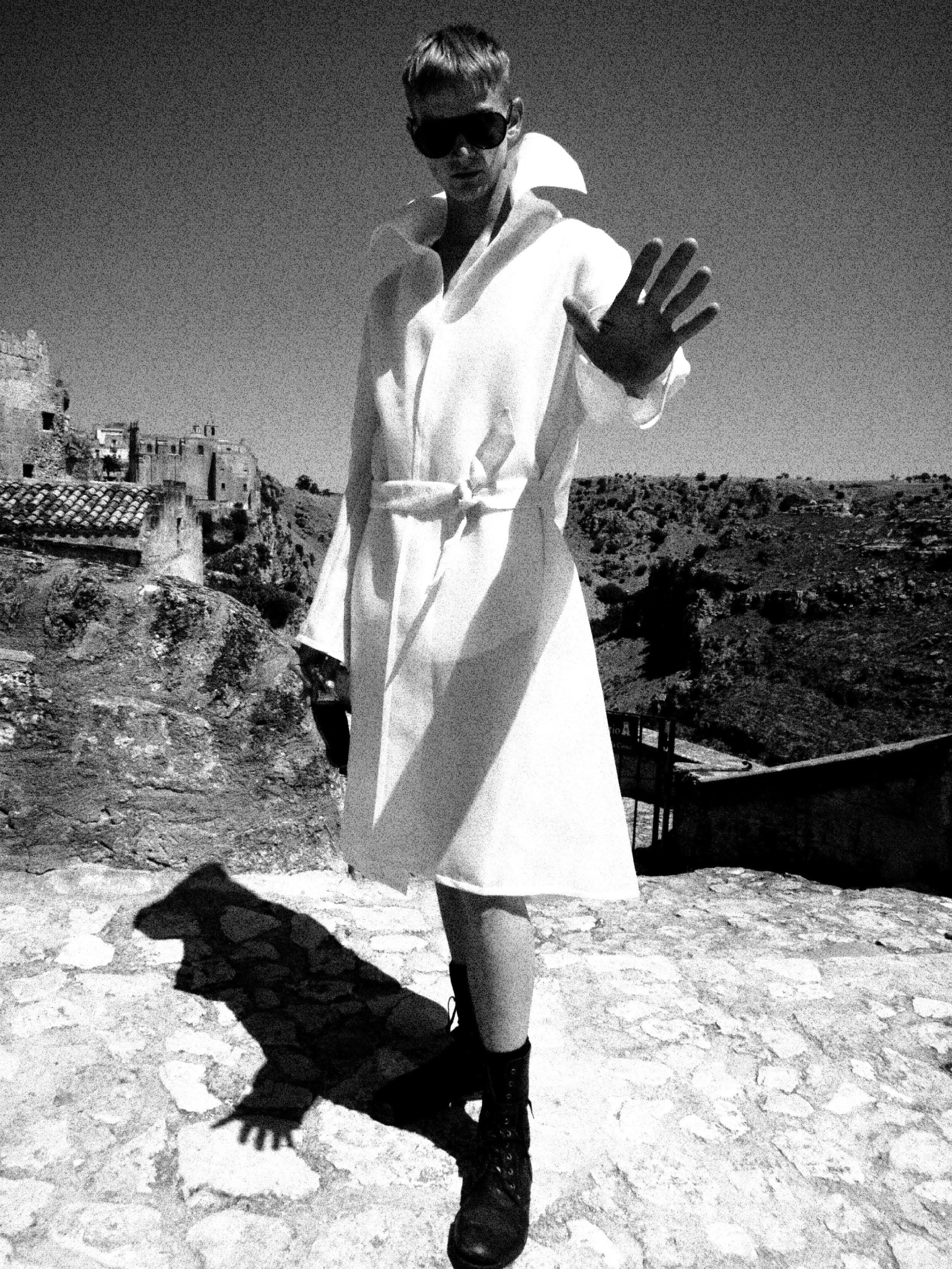 A person wearing a white coat, black boots, and sunglasses standing on rocky terrain with a modern landscape in the background. The person is making a gesture with their hand.