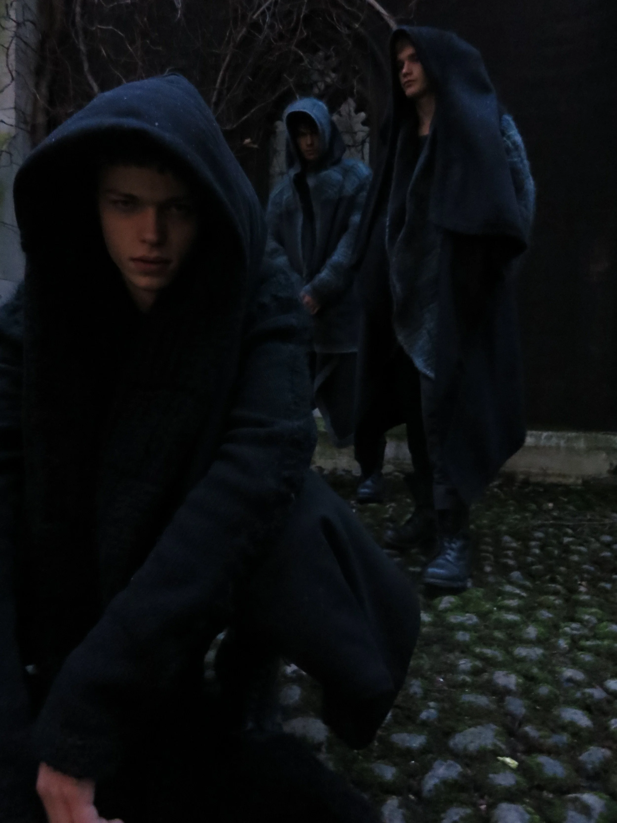 Three young men wearing dark hooded jackets standing outdoors at night, with one in the foreground leaning forward and two in the background standing close together.