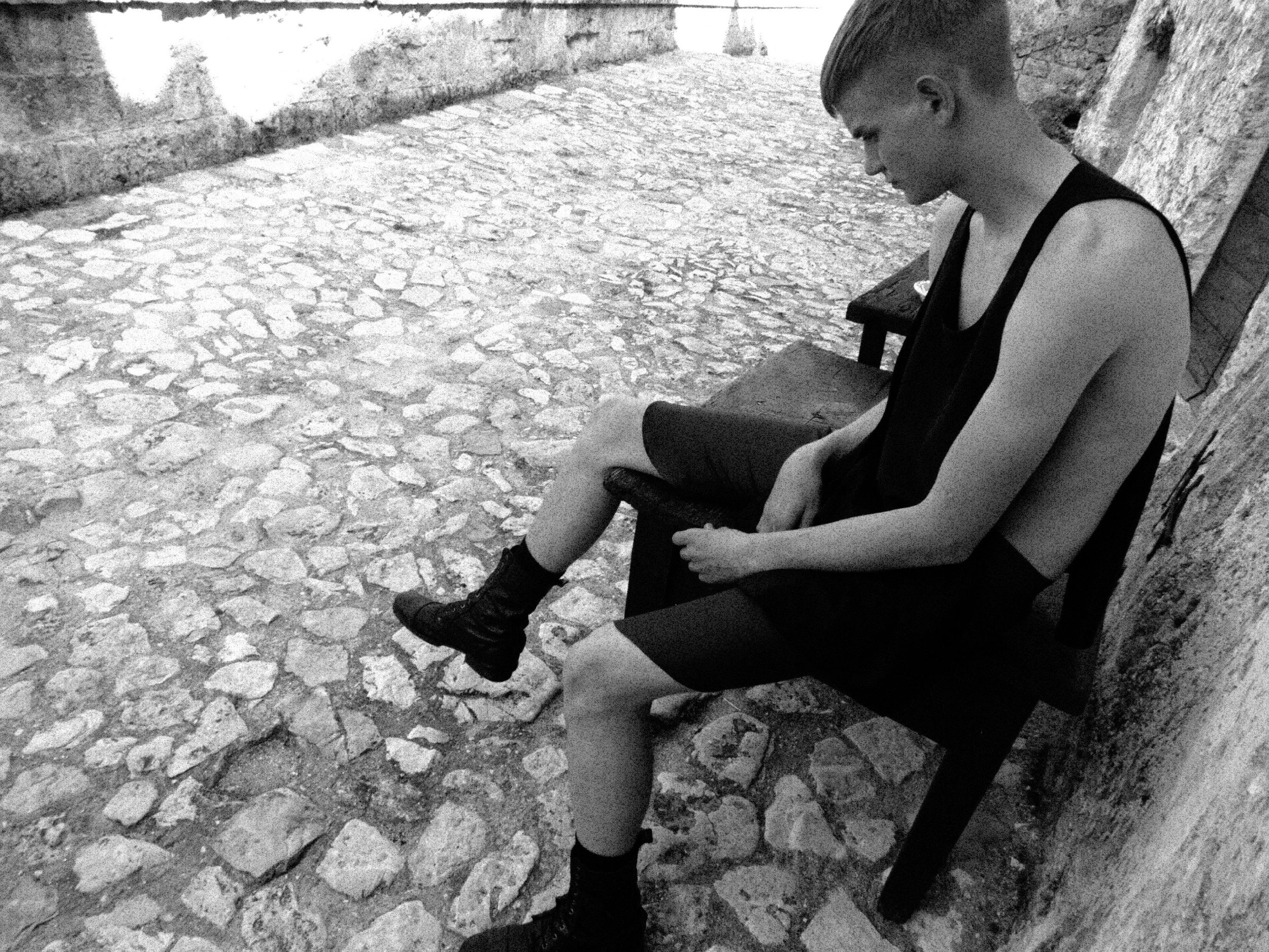 Black and white photo of a young woman sitting on a bench with her head bowed and eyes closed, wearing a sleeveless top, skirt, and boots, against a stone and brick background.