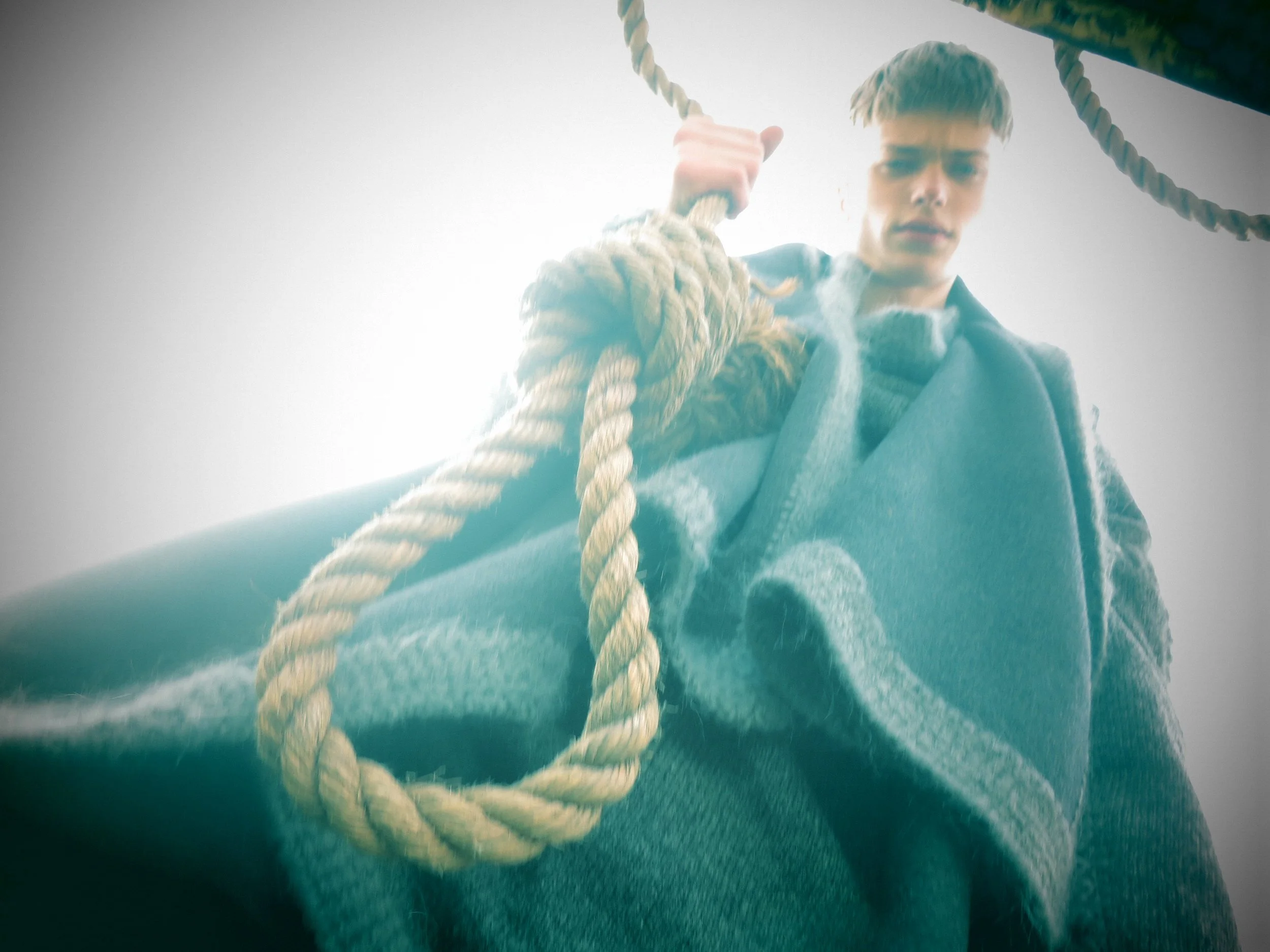 A young man with short hair holding onto a thick rope while standing on a boat, with a cloudy sky in the background.