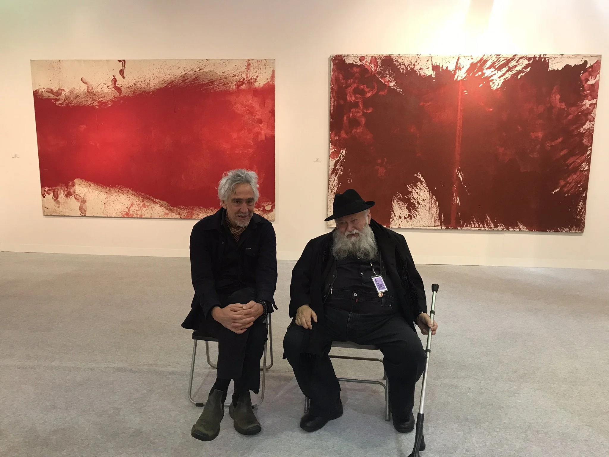 Two men sitting on chairs in an art gallery with large red abstract paintings behind them. The man on the right has a long gray beard, is wearing a black beanie, and holds a cane. The man on the left has gray hair and is wearing dark clothing.