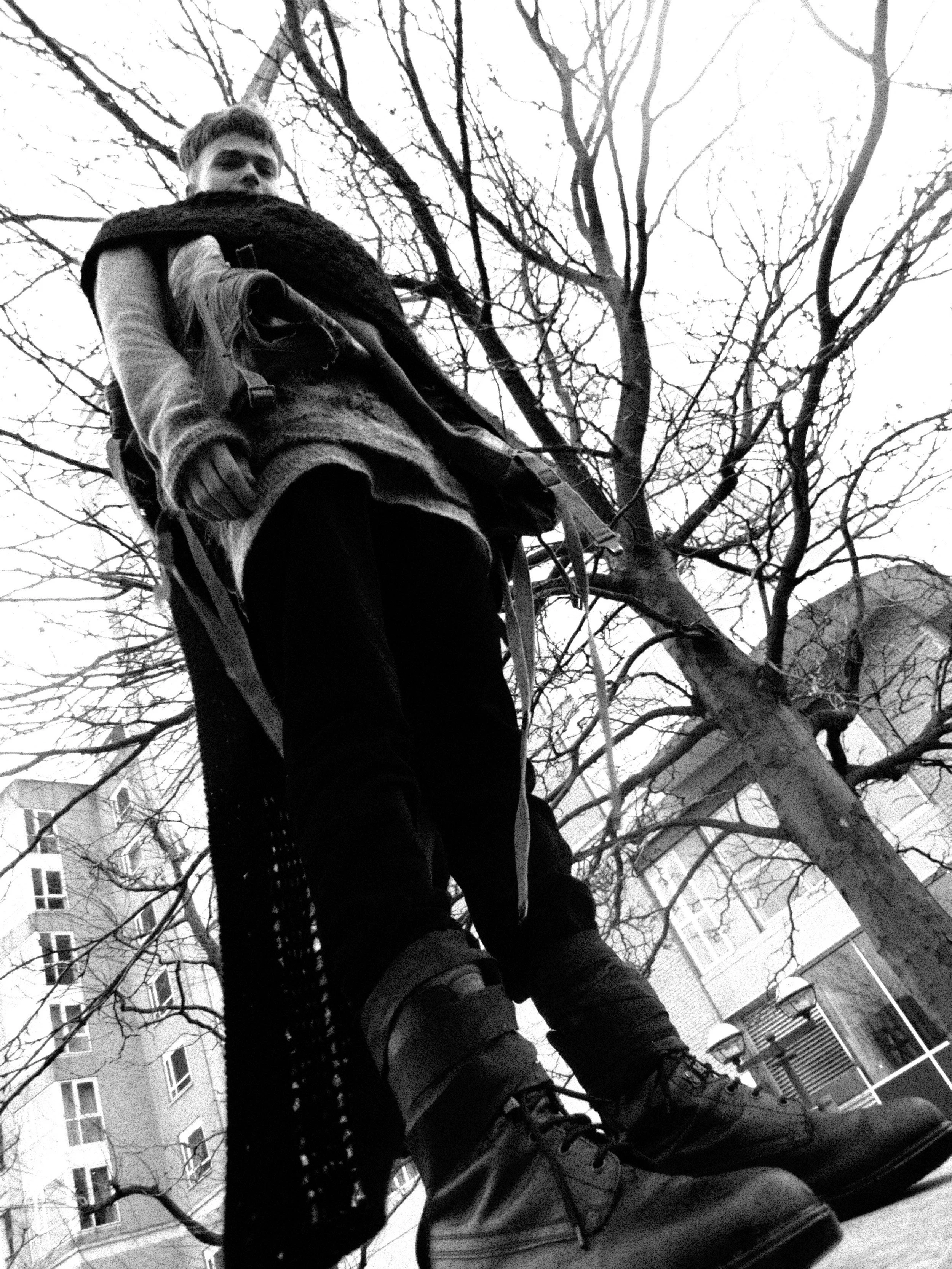 A person wearing winter clothing, shoes, and a backpack is standing outdoors among leafless trees, with buildings and a fence in the background. The photo is taken from a low angle looking up.