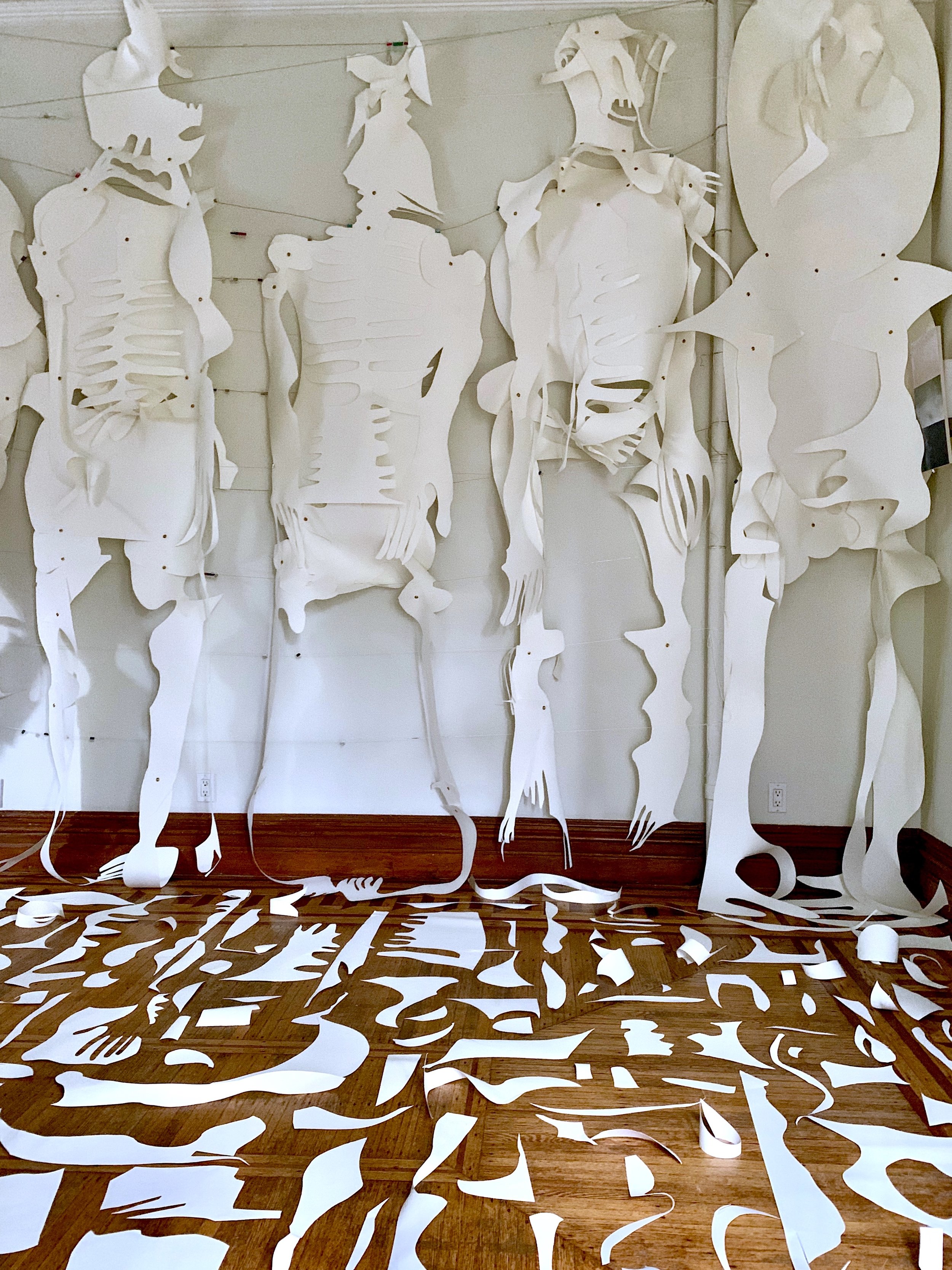Installation art featuring paper cutouts of human skeletons and shapes, with loose paper pieces on a hardwood floor.