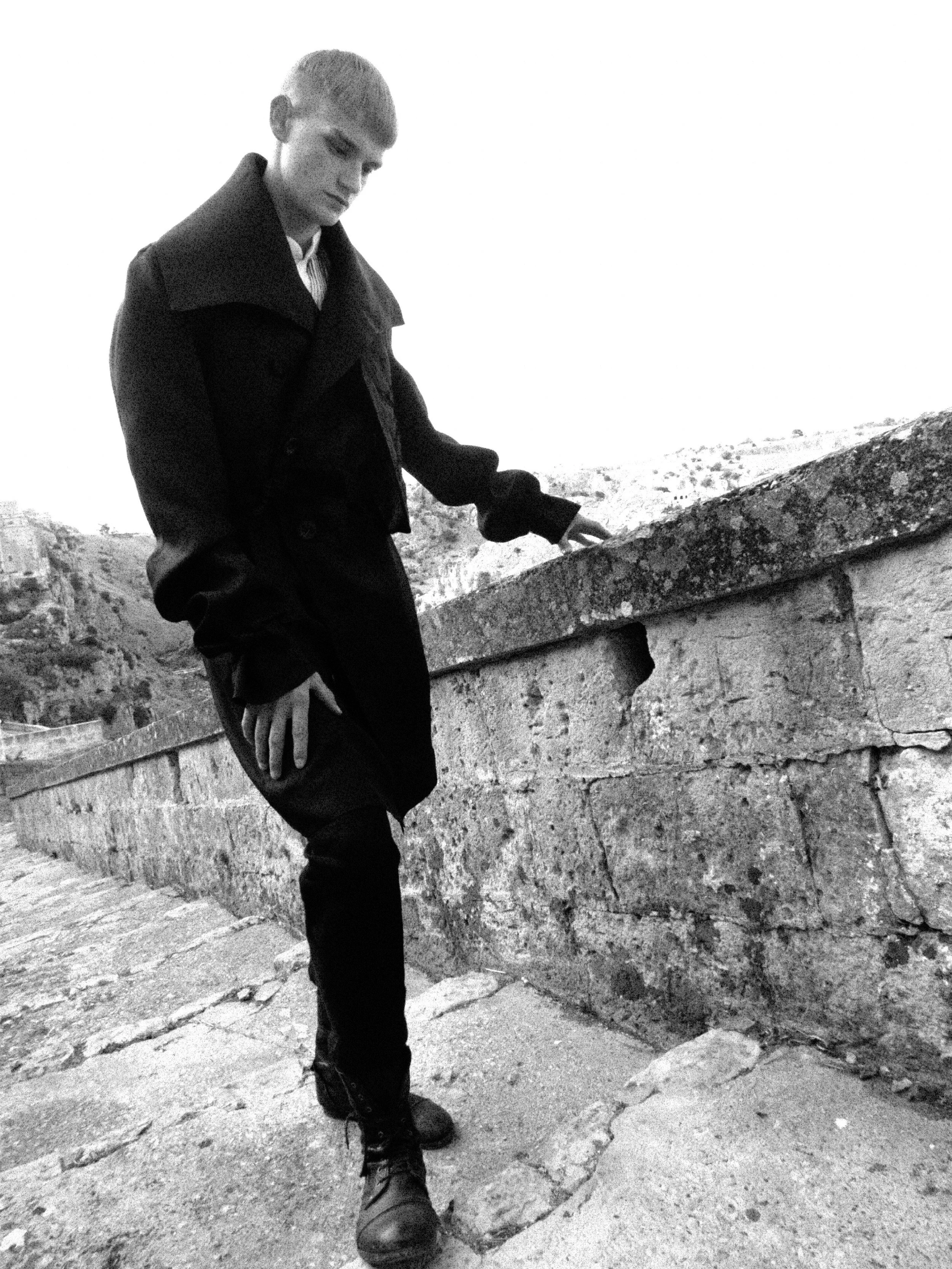 A person standing outdoors on a stone staircase, wearing a dark jacket and boots, holding onto the stone railing with one hand, with a blurry landscape in the background.