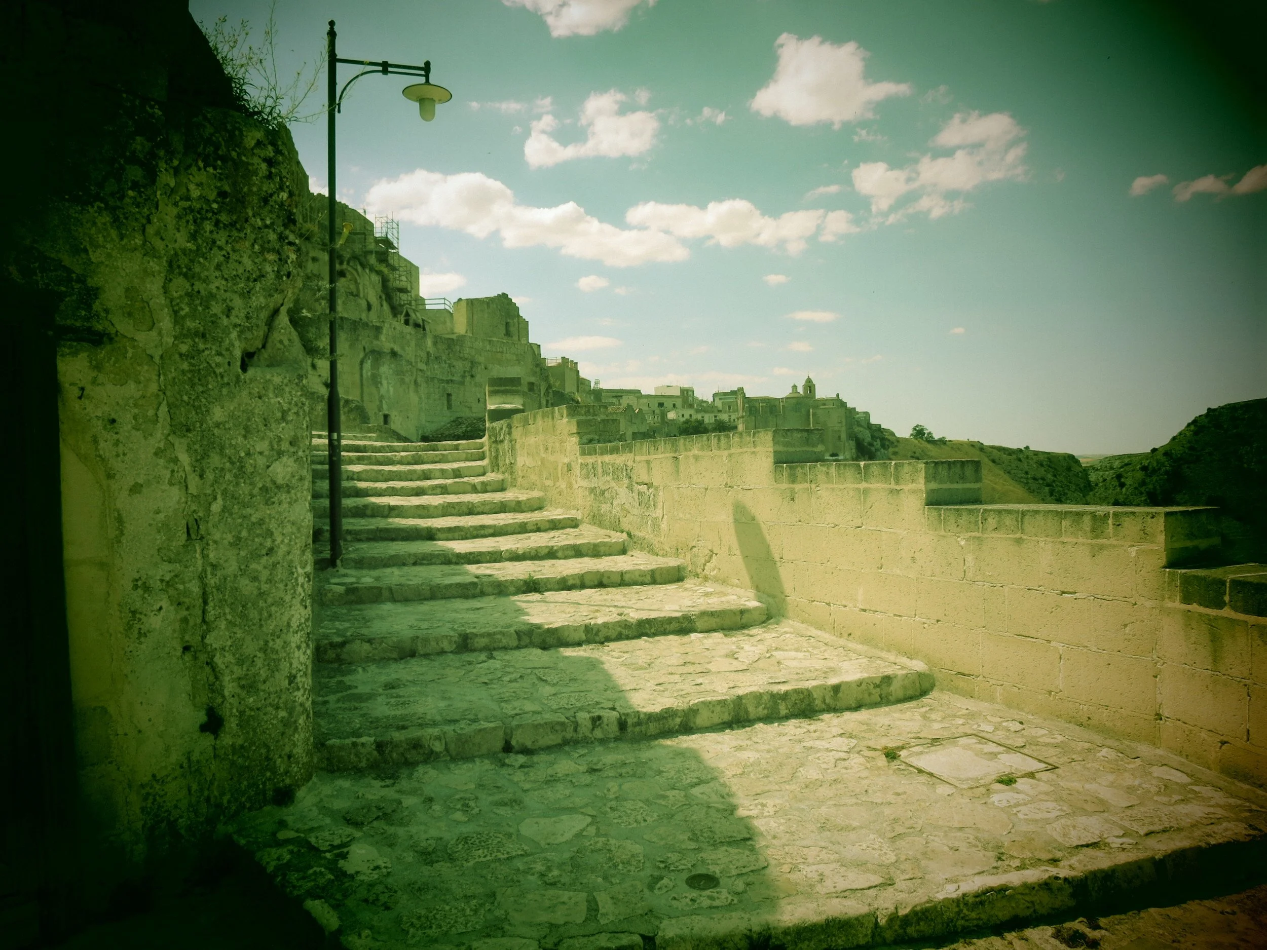 Stone stairs leading up to an ancient stone fortress or castle under a partly cloudy blue sky. A streetlamp is visible on the left side of the image.