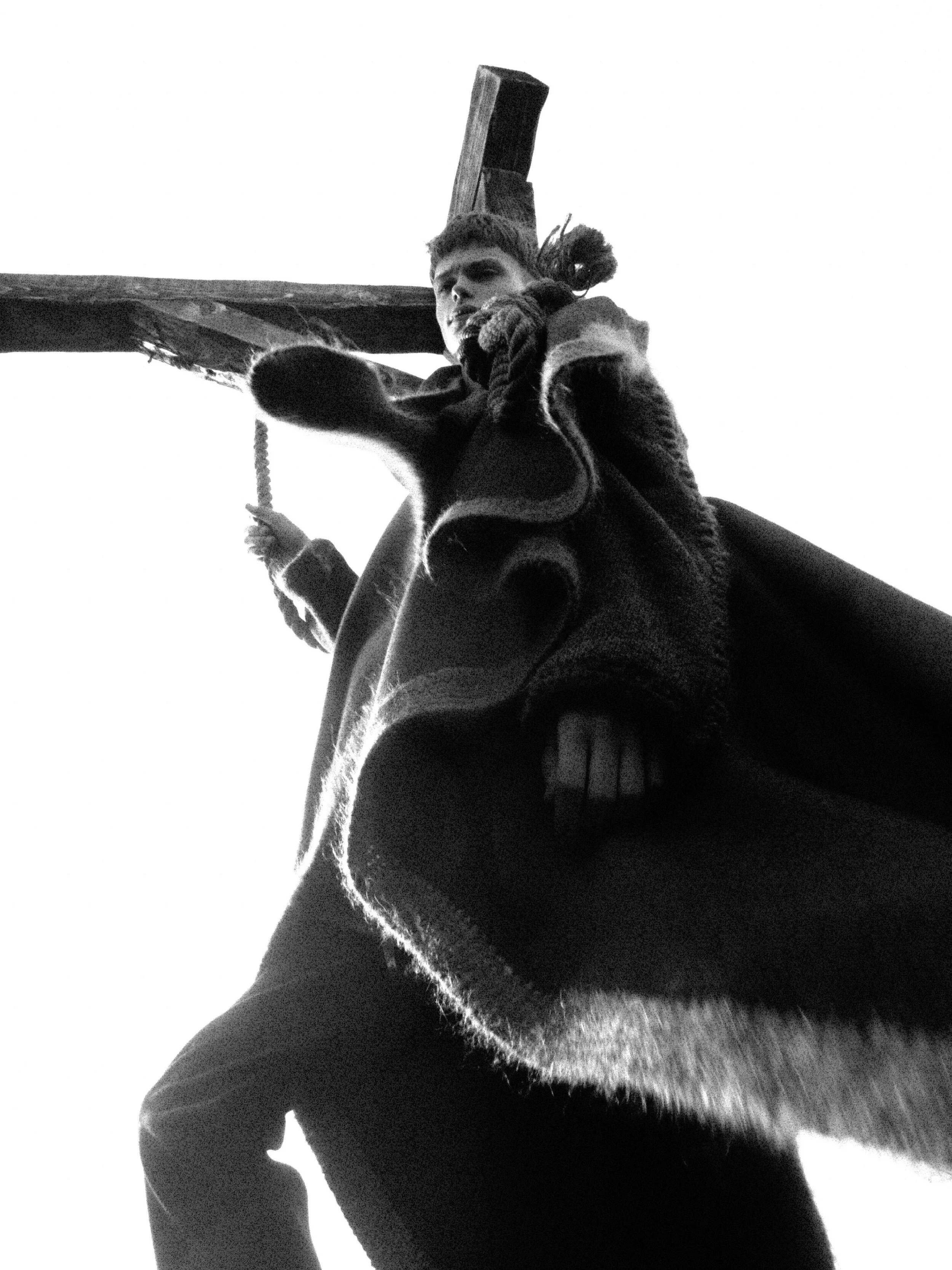 Low angle black-and-white photo of a person carrying a large wooden cross, with a dog in the foreground.