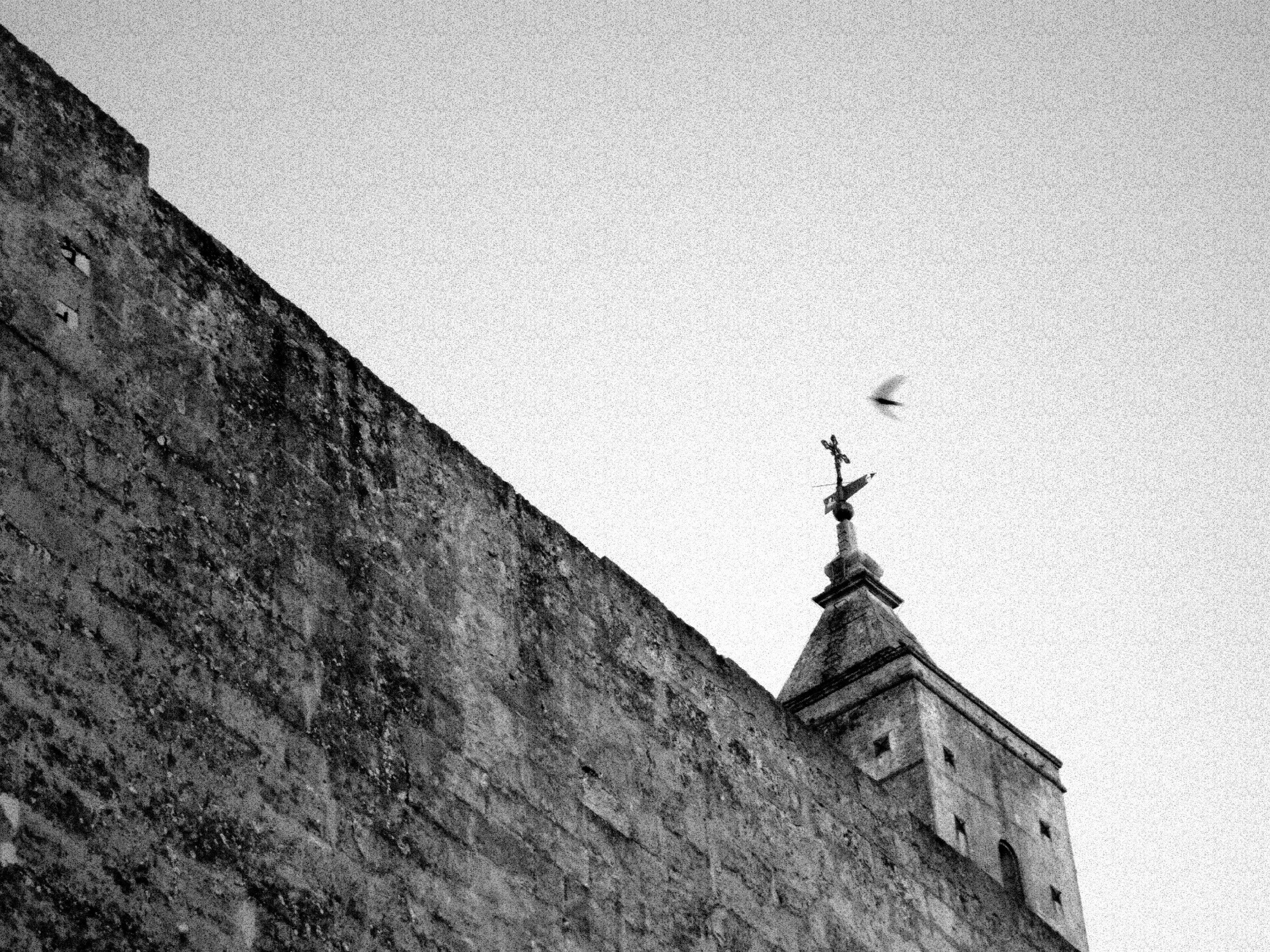 A black and white photo of a church or cathedral with a cross on top, taken from a low angle. The photograph features a textured stone wall in the foreground and the building extending upward with a weather vane or similar decorative element at the p