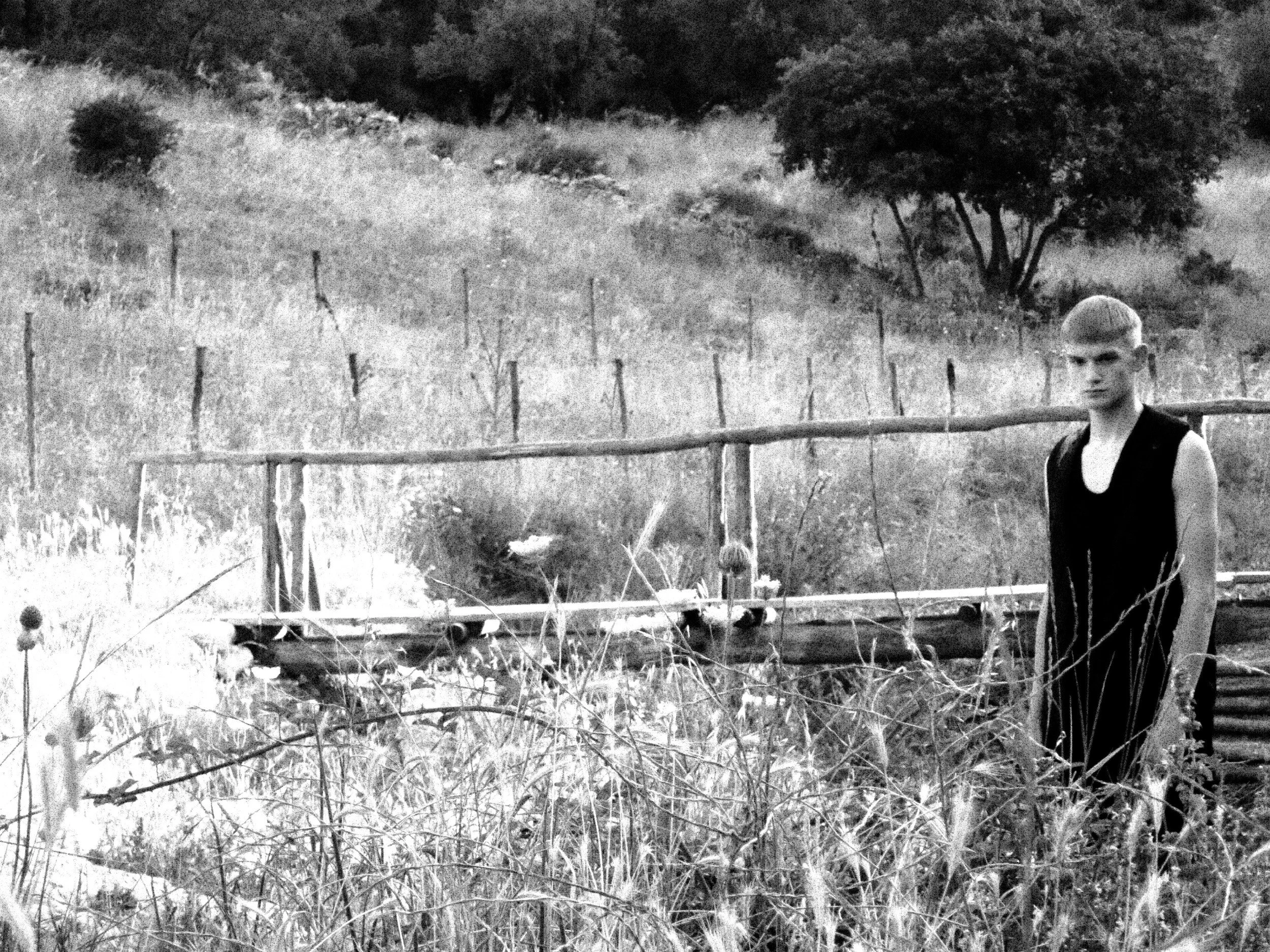 A black and white photo of a young man with short hair standing outdoors among tall grass and wild vegetation, with a fence in the background and trees on a hillside.