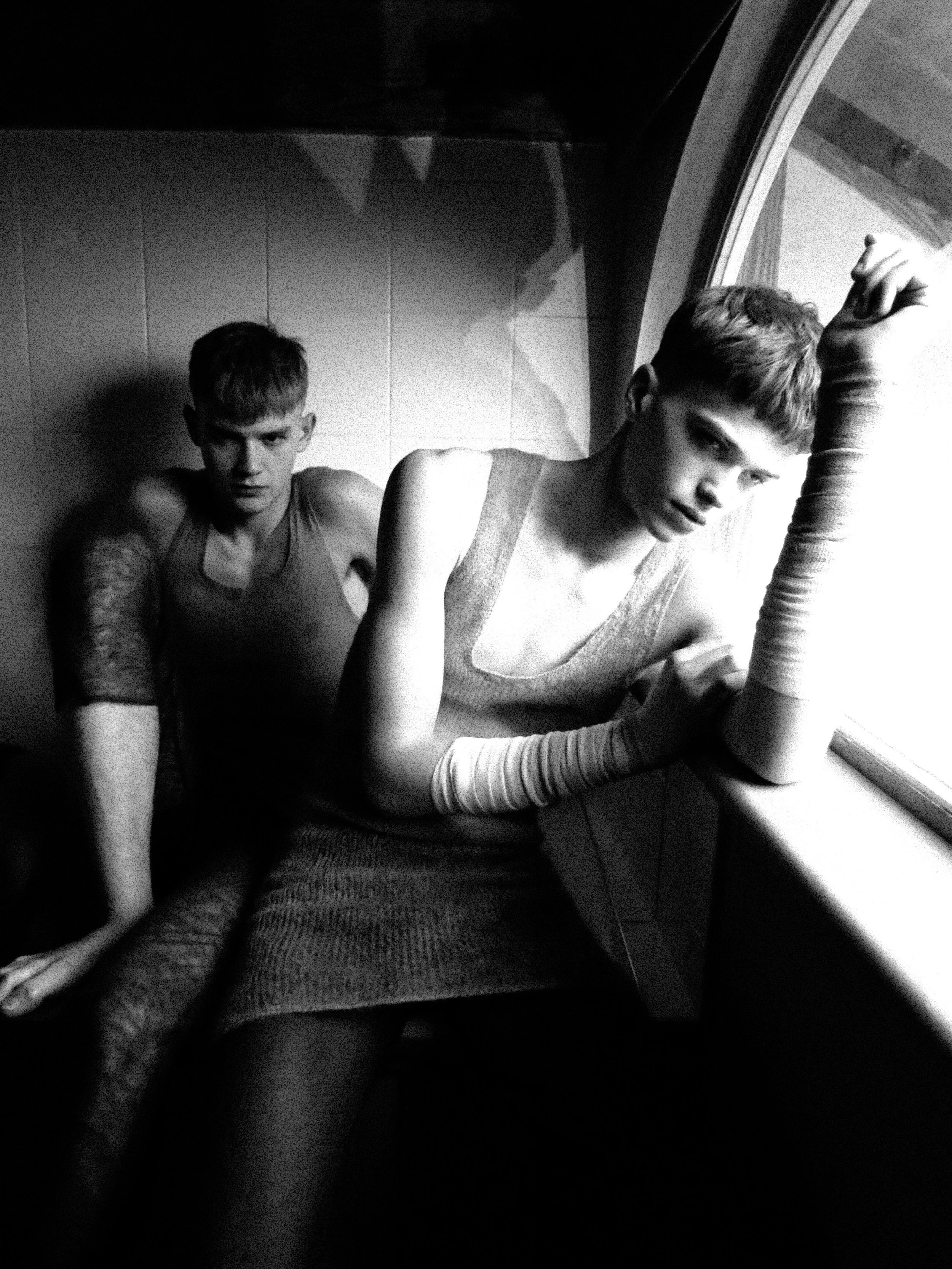 Black and white photo of two young men sitting on the floor near a window, one resting his arm on his knee and the other leaning against the wall with a serious expression.