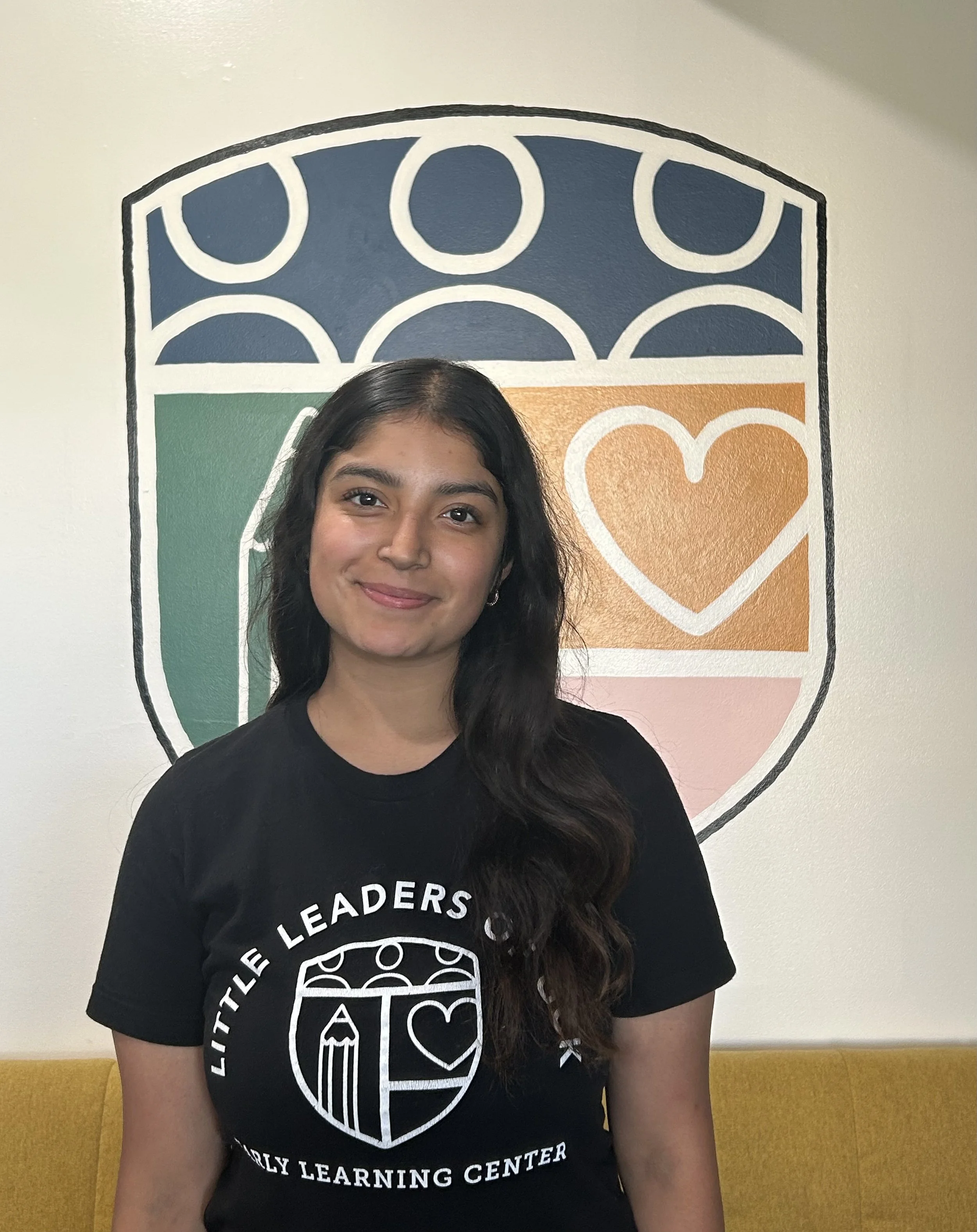 A smiling young woman with long dark hair, wearing a black T-shirt with a circular logo. She is standing in front of a colorful wall mural with a shield design, a heart, and geometric shapes.