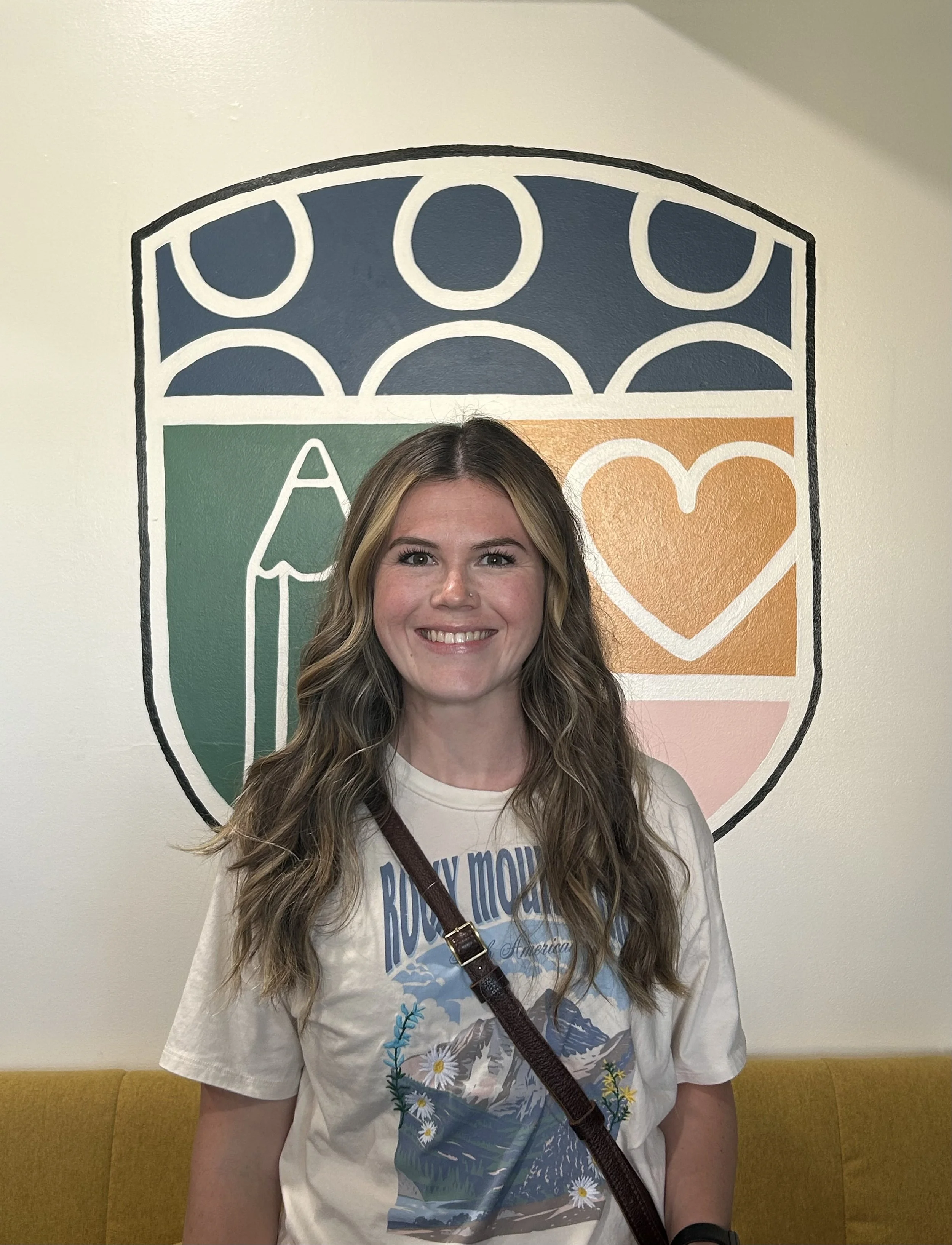 A young woman with long wavy hair smiling, wearing a white T-shirt with a mountain and floral design, standing in front of a decorative mural on a wall featuring a shield with various symbols including a leaf, heart, and umbrella.