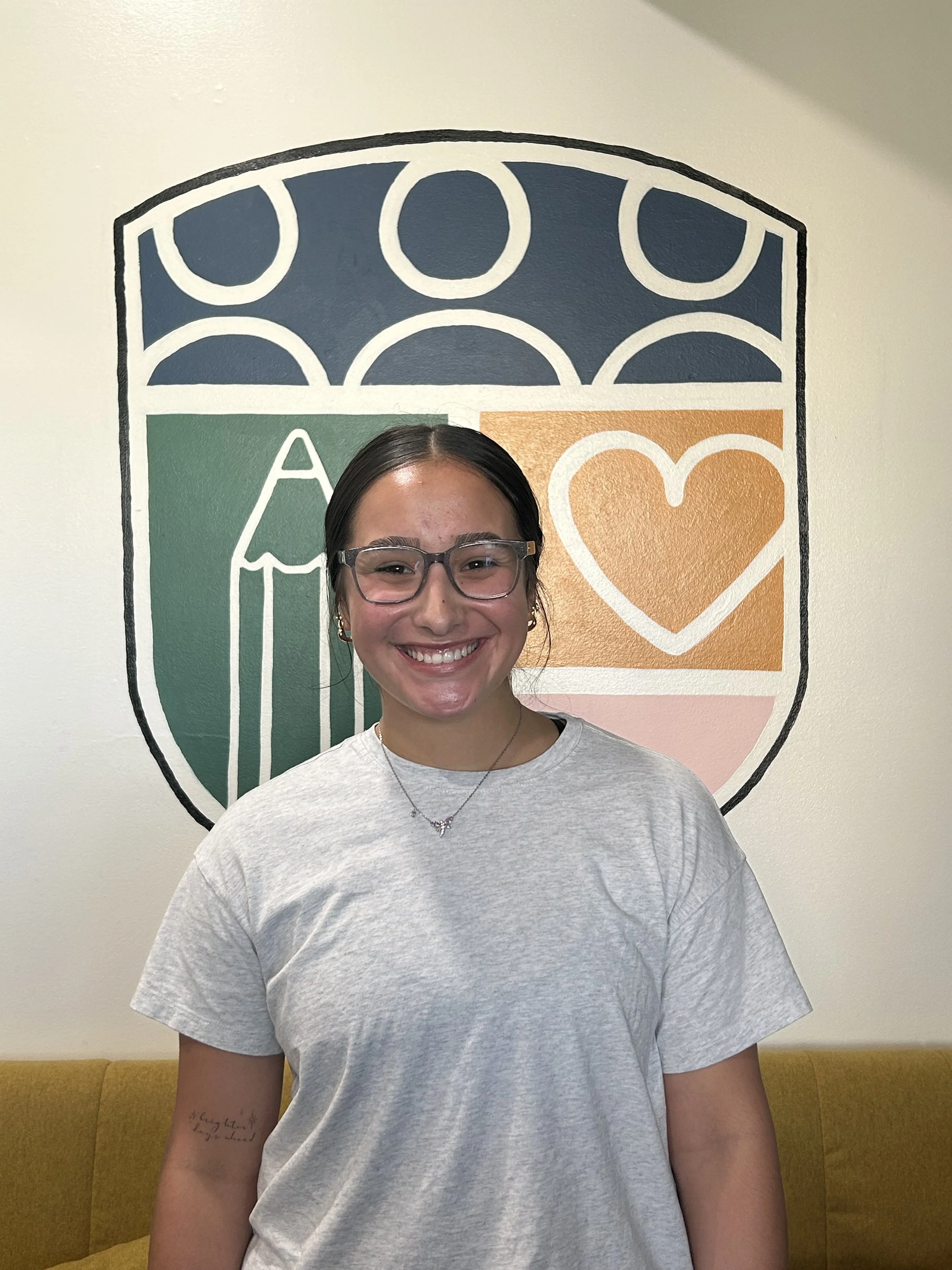 A young woman with glasses and dark hair smiling in front of a wall art with a paint vault and a heart symbol.