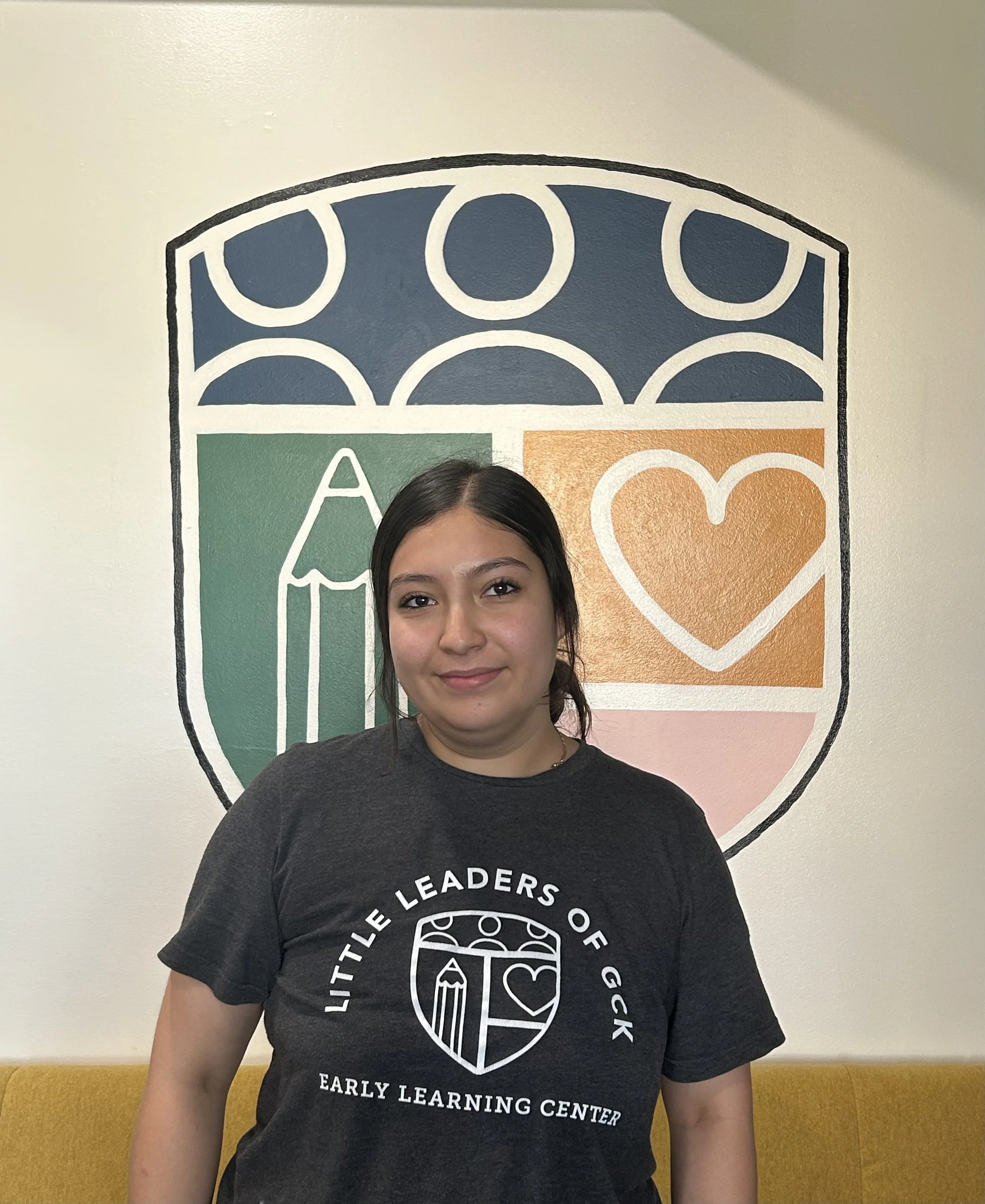 A young girl with dark hair in pigtails, wearing a black T-shirt with white text and graphics, is standing in front of a colorful mural on a wall. The mural features abstract shapes including a large orange heart and various blue, green, and pink sections.