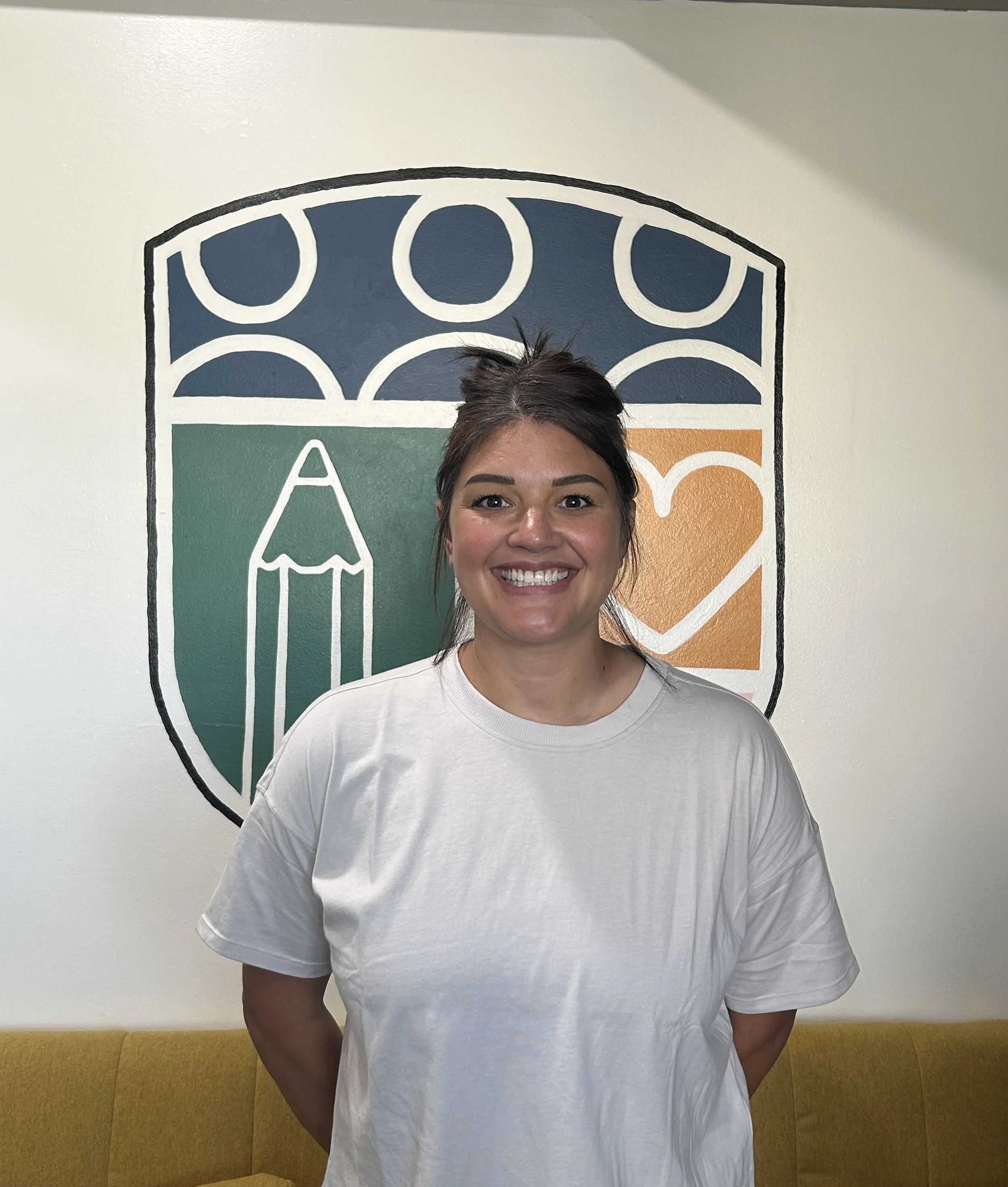 A woman with dark hair, tied into a bun, smiling at the camera, standing in front of a wall with a colorful crest or logo that features a pencil and shield design.