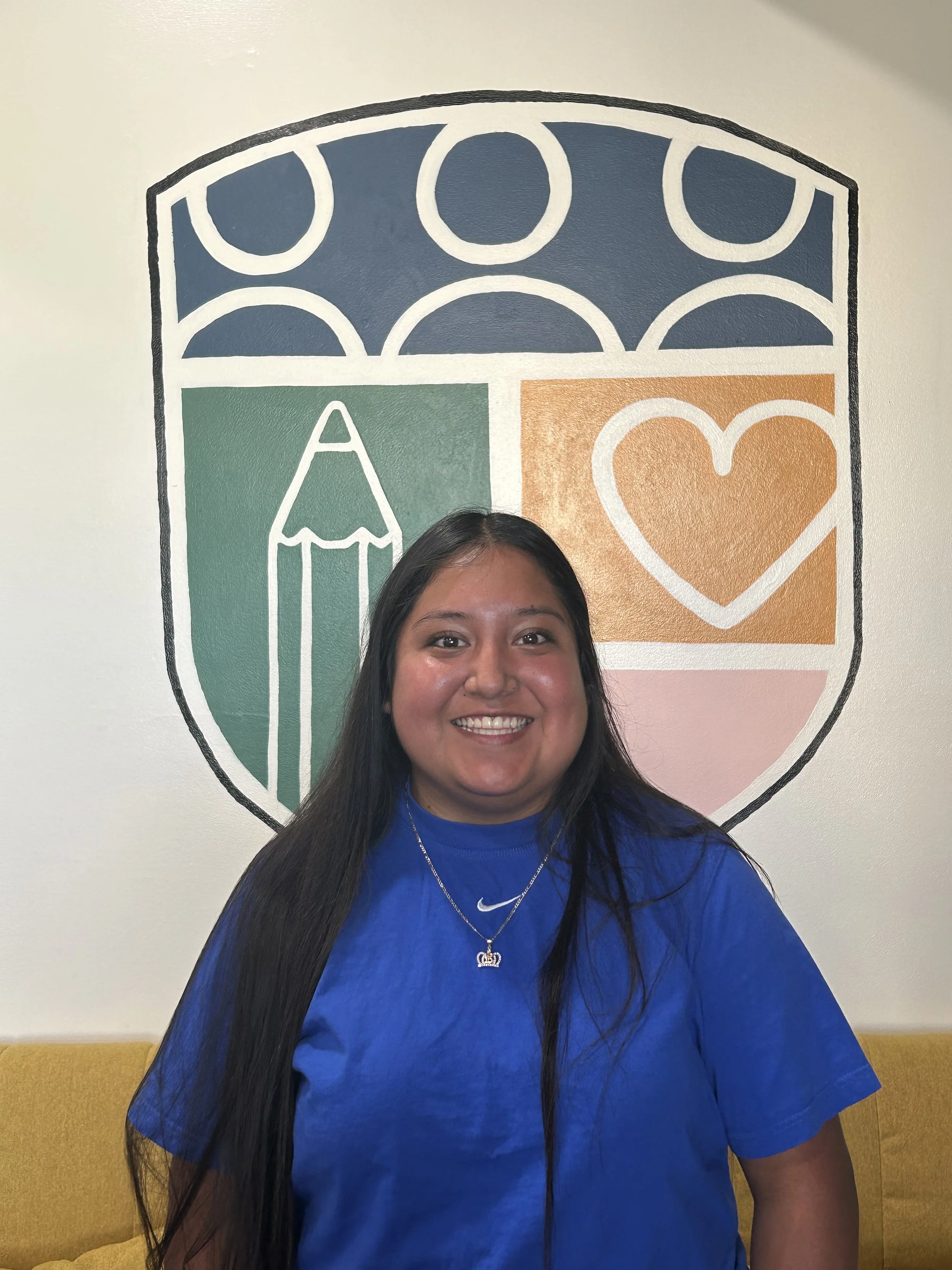 A woman with long black hair wearing a blue Nike shirt and a silver necklace smiling in front of a colorful wall mural with a pencil, a heart, and abstract shapes.