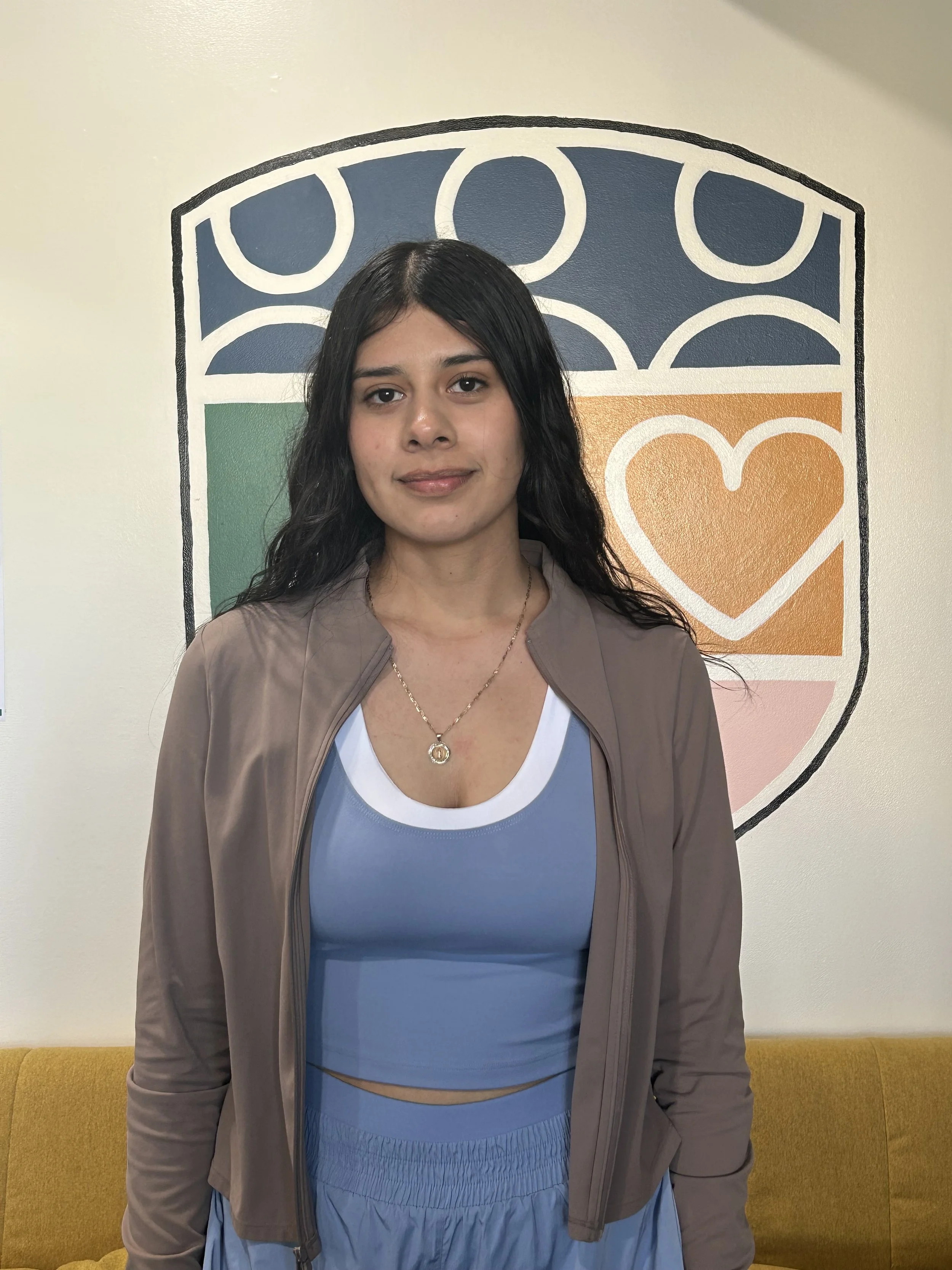 A young woman with dark hair standing in front of a colorful mural featuring geometric shapes and a heart design.