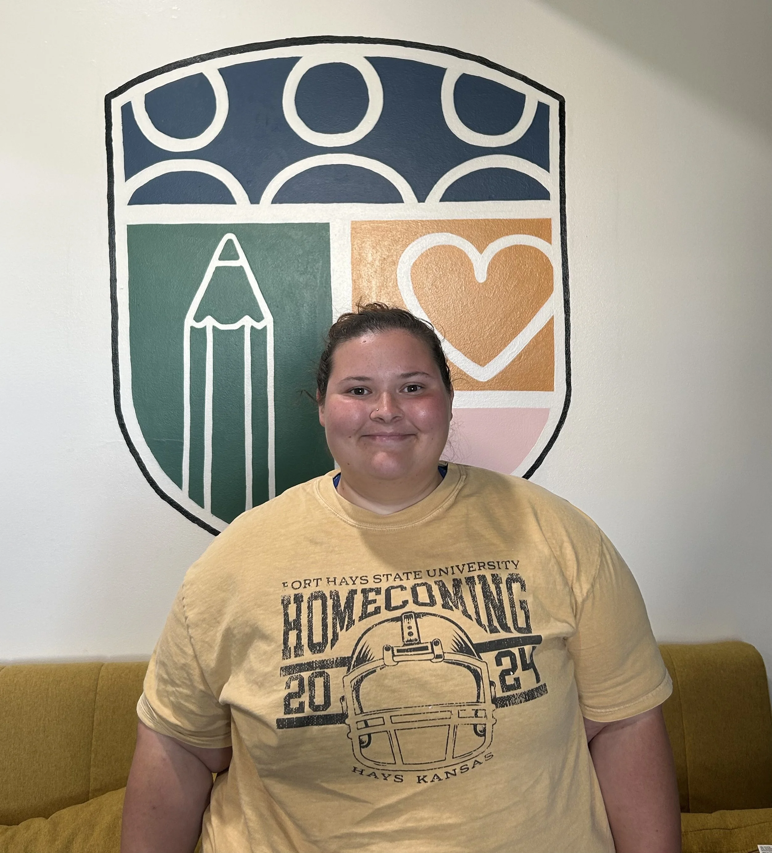 A person smiling, wearing a yellow T-shirt with a graphic design of a toolbox and text that reads "Hays Kansas," in front of a wall with a large colorful mural featuring abstract shapes and symbols, including a heart, a pencil, and other geometric figures.