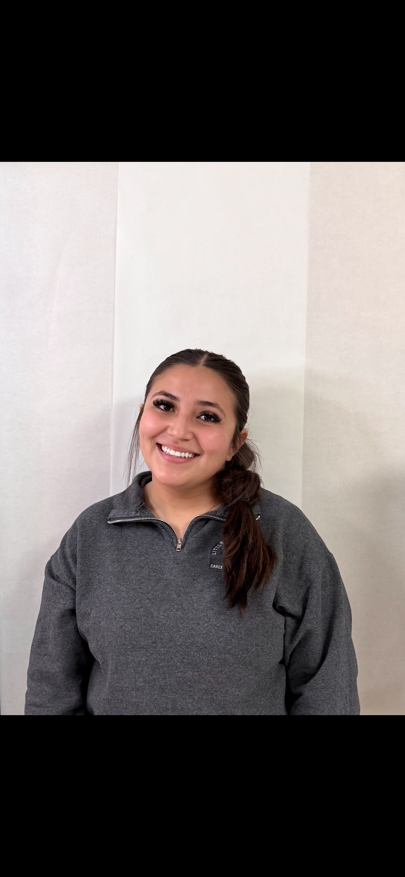 A smiling young woman with long brown hair in a side braid, wearing a gray zip-up sweater, standing against a plain white wall.