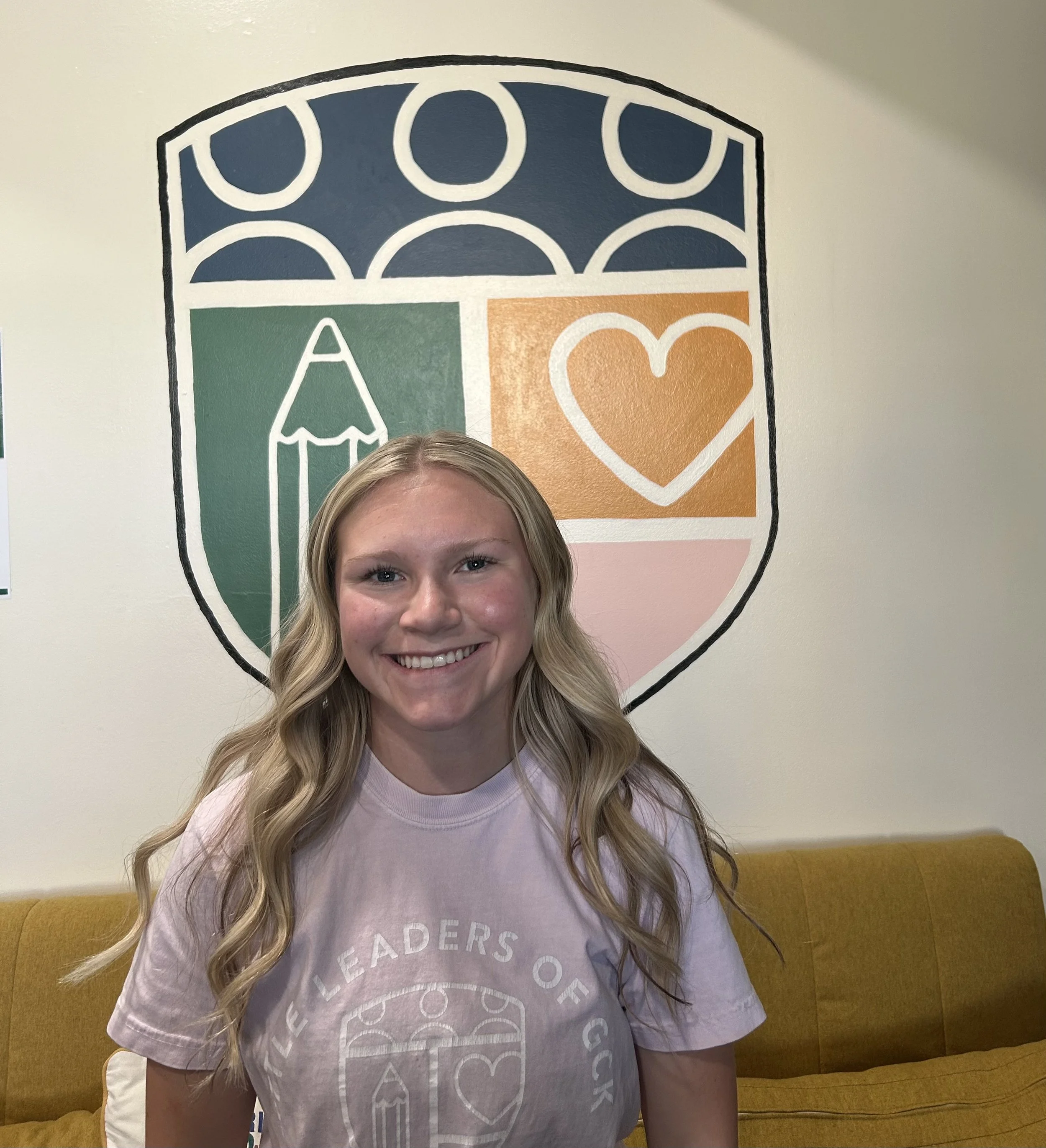 Smiling young woman with long blonde hair sitting on a yellow upholstered bench in front of a mural with abstract shapes and icons in blue, green, orange, and pink colors.
