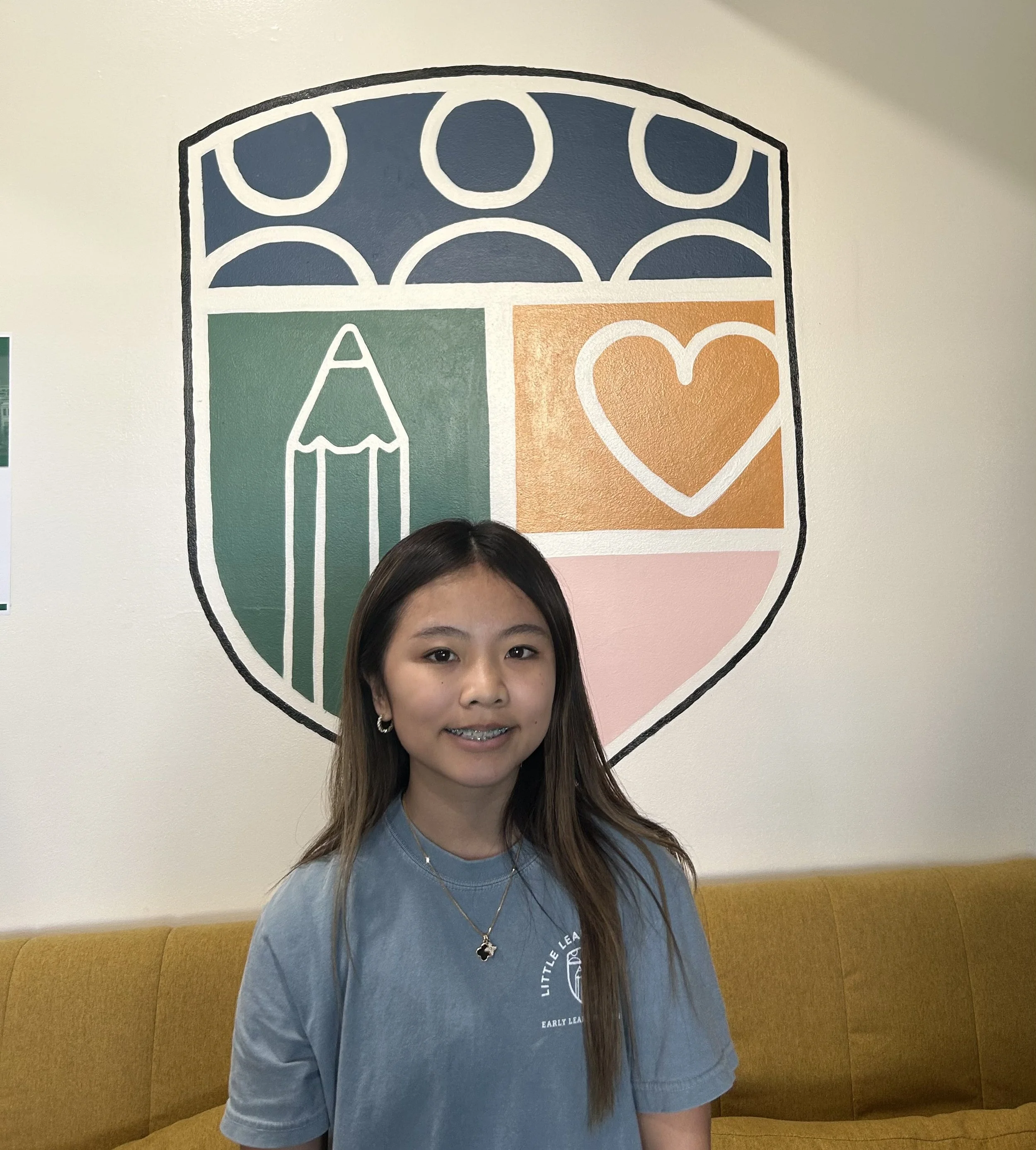 A young woman with long hair is standing in front of a colorful wall mural featuring a simple, stylized design of overlapping circles and abstract shapes. The mural includes a section with a green fish shape and an orange heart shape. She is wearing a blue T-shirt with a circular white logo and a star necklace.