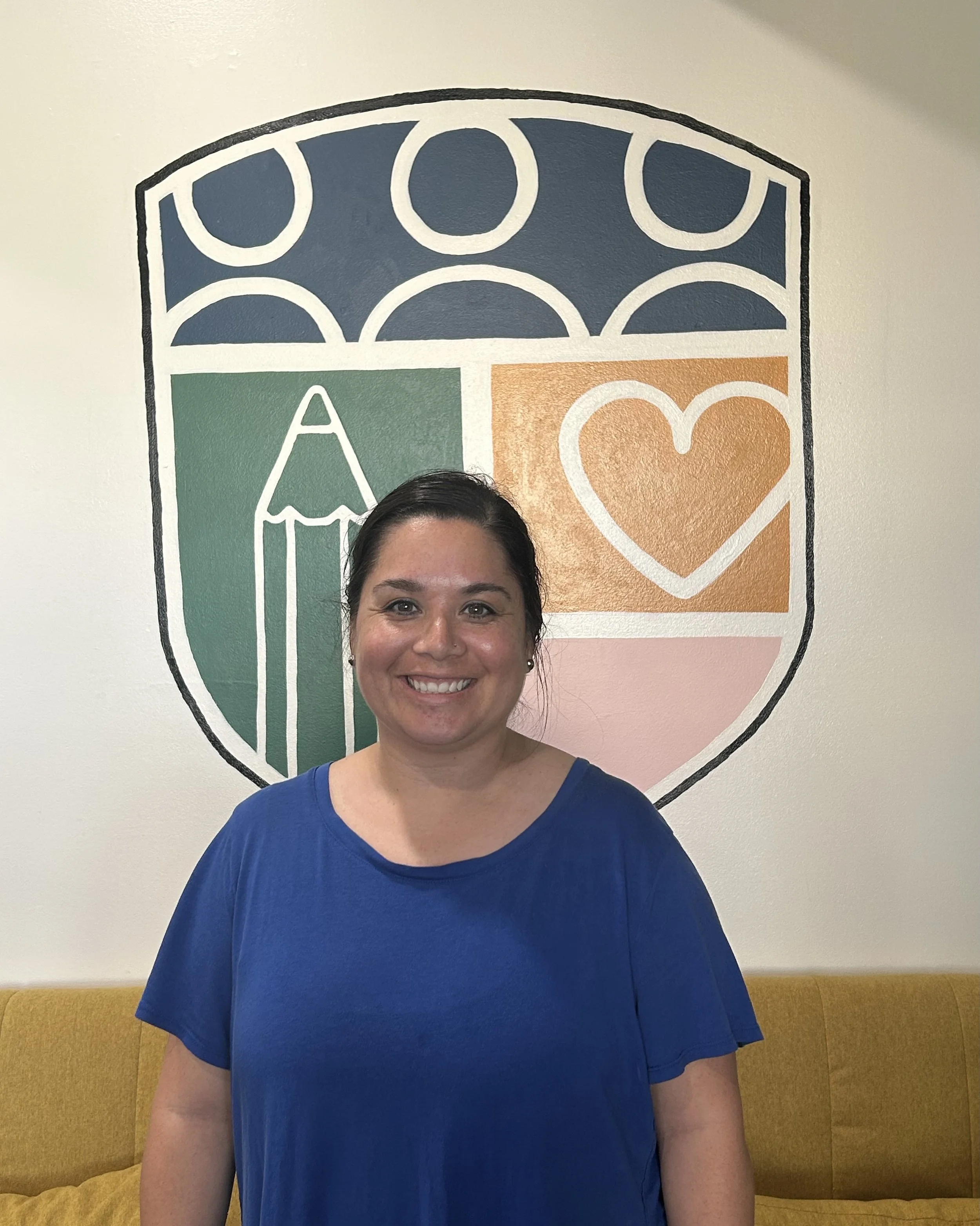 A woman with dark hair, smiling, wearing a blue shirt, standing in front of a colorful wall art featuring a shield with symbols including a pencil, a heart, and circles.
