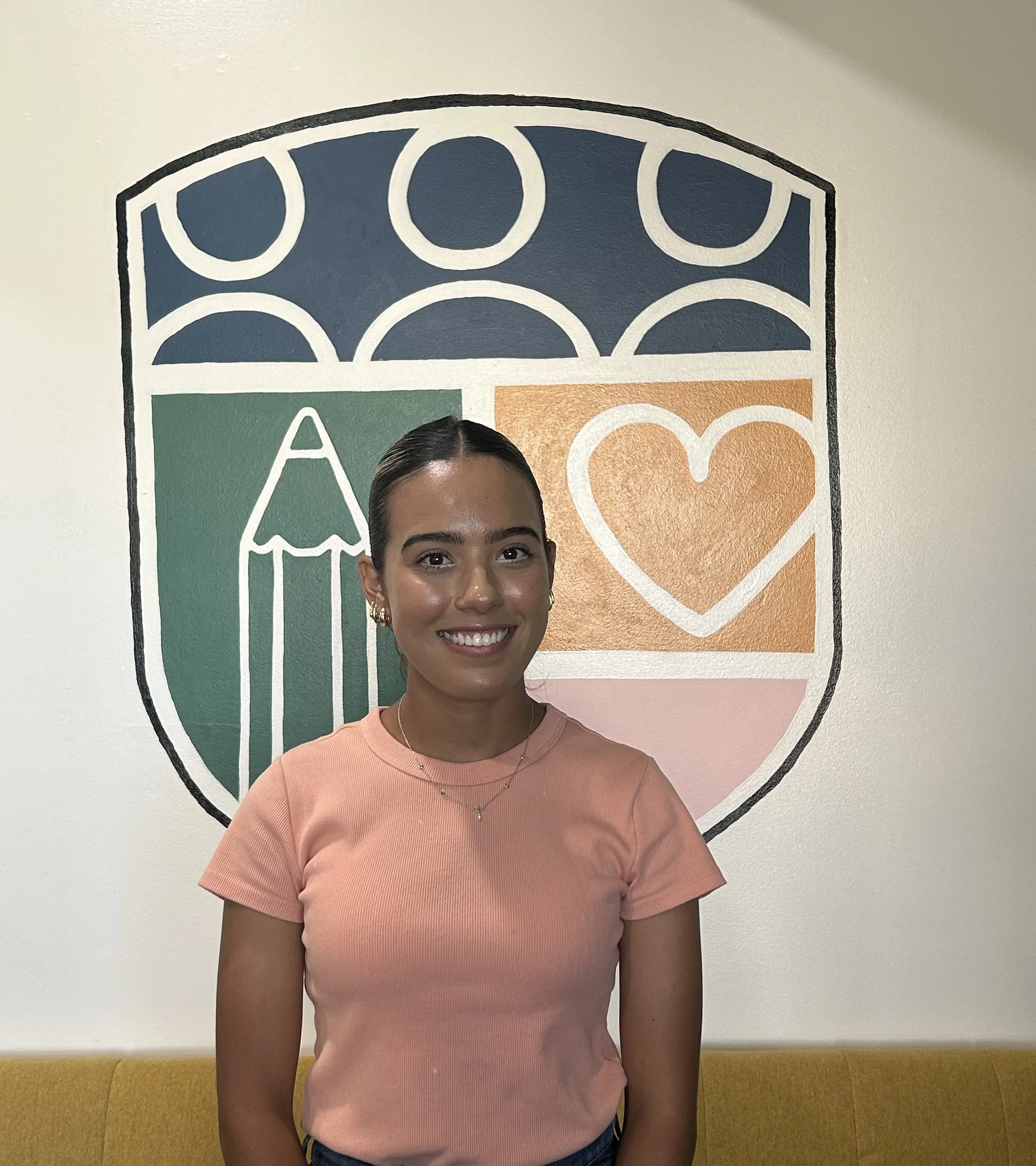 Young woman with dark hair, smiling, wearing a peach-colored T-shirt and jewelry, standing indoors in front of a colorful wall with a geometric design and text.