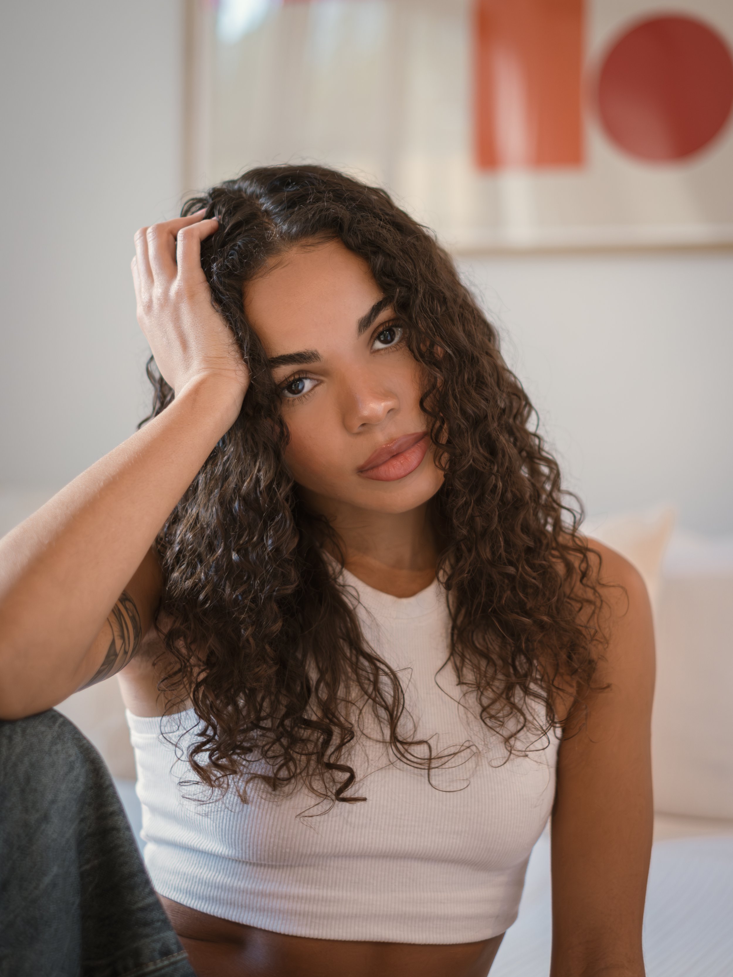 A model with curly hair, wearing a white sleeveless top, sitting indoors with a neutral expression, touching her head with her left hand.