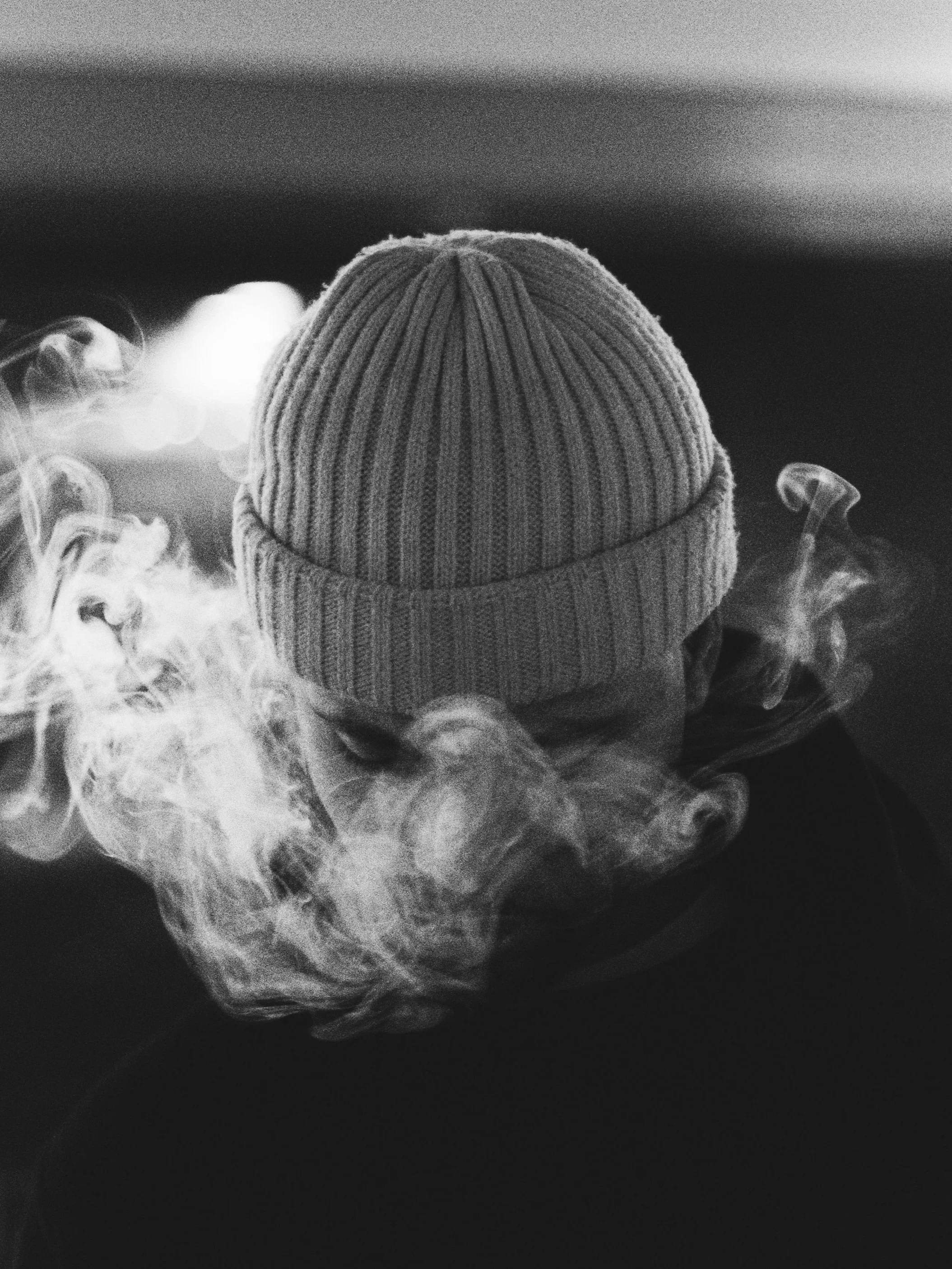 A person wearing a knit beanie hat, with their head slightly bowed, surrounded by swirling smoke or vapor, in black and white.